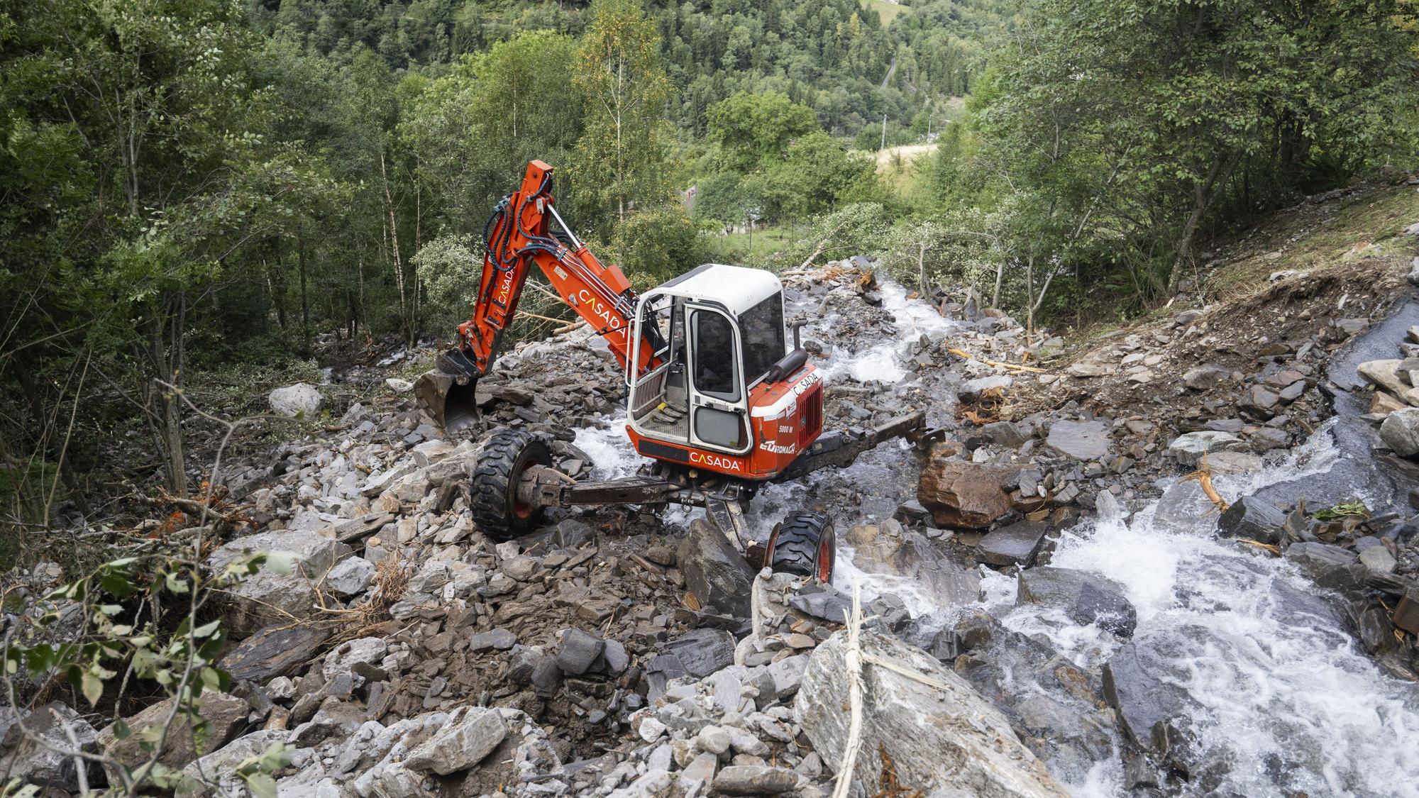 Ein orangeroter Bagger auf einem Abhang voller Geröll. Ein Bach fließt vorbei.