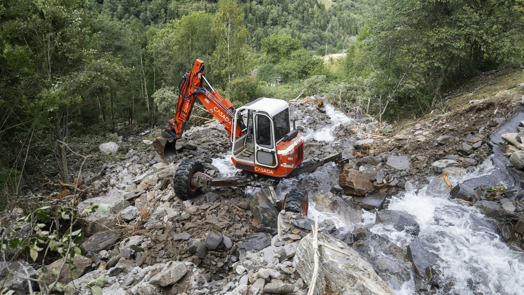 Ein orangeroter Bagger auf einem Abhang voller Geröll. Ein Bach fließt vorbei.