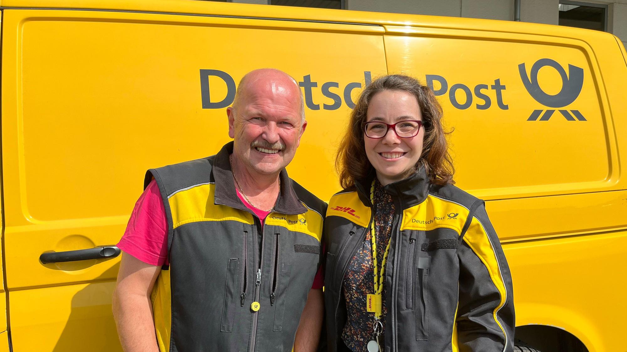 Ein Mann mit Glatze und Schnurrbart steht vor einem gelben Kleinbus mit der Aufschrift Deutsche Post. Neben ihm steht eine Frau mit Brille und einer schwarzen Jacke mit gelben Einsätzen.