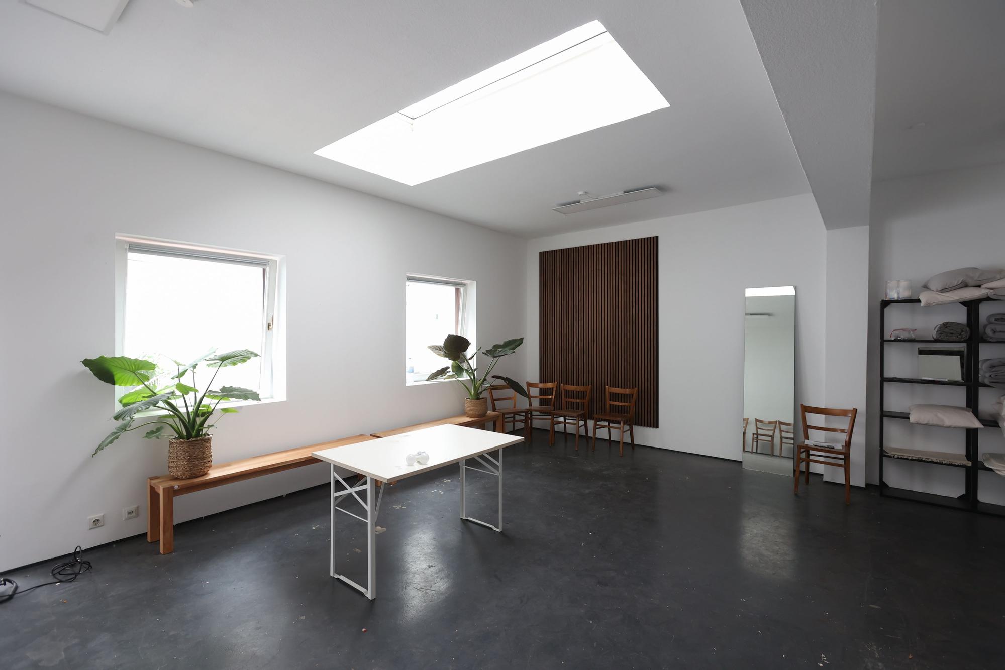 Ein heller Raum mit großen Fenstern und weißen Wänden. Eine Bank, Stühle und ein Spiegel stehen an der Wand.