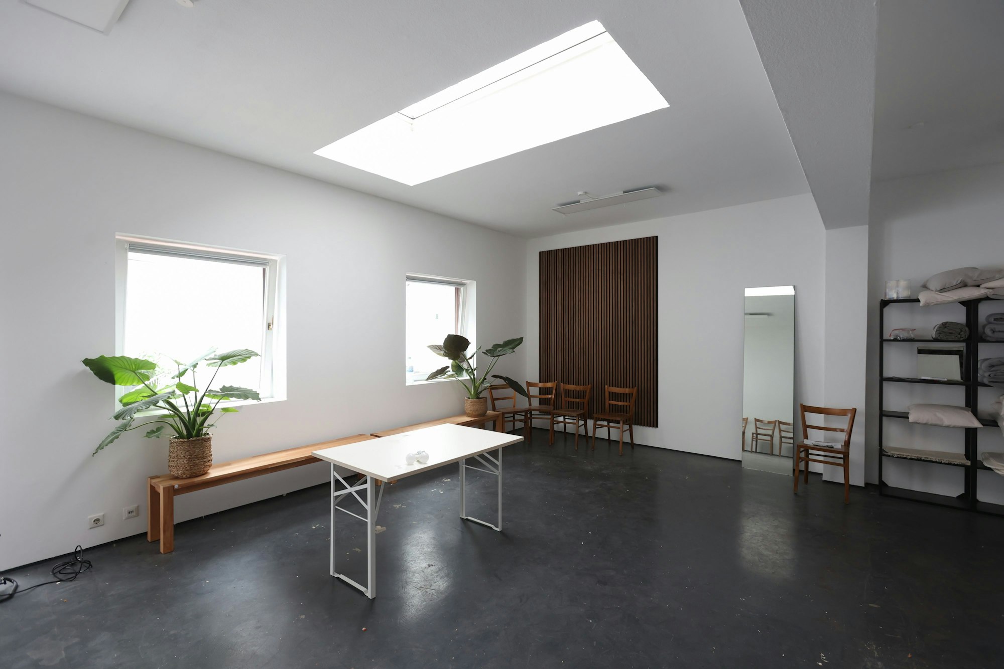Ein heller Raum mit großen Fenstern und weißen Wänden. Eine Bank, Stühle und ein Spiegel stehen an der Wand.