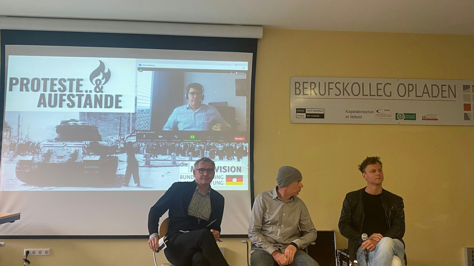 Podiumsdiskussion zu Aufständen und Protesten am Berufskolleg Opladen: Dr. Günter Hinken (v.l.), Tim Eisenlohr, Alex Wernke, auf der Leinwand: Ehsan Ayatollahi.