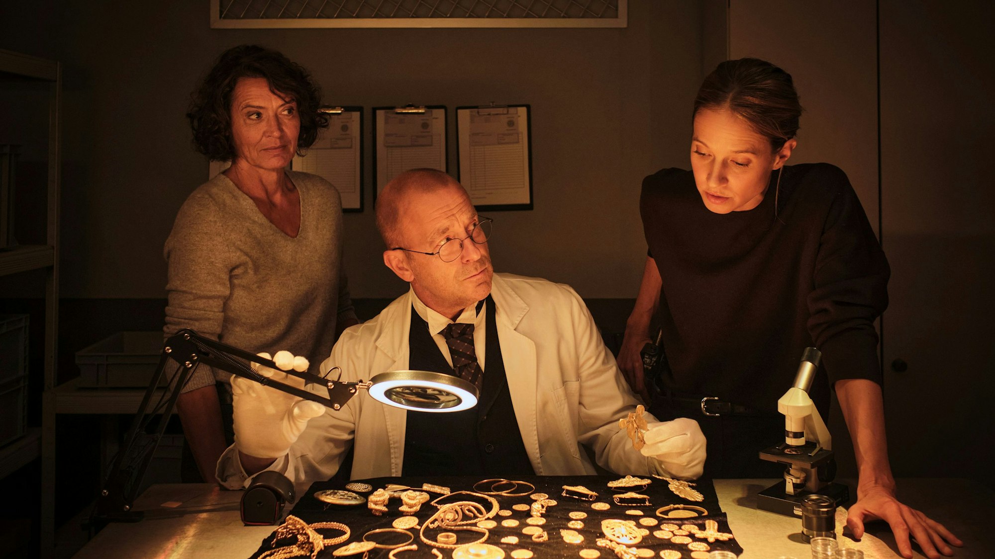 Johanna Stern beugt sich über den Goldschatz, der auf einer samtenen Decke liegt. Der Kurator Albert Dürr sitzt vor dem Schatz und beäugt Stern kritisch. Auch Odenthal schaut skeptisch zu ihrer Kollegin.