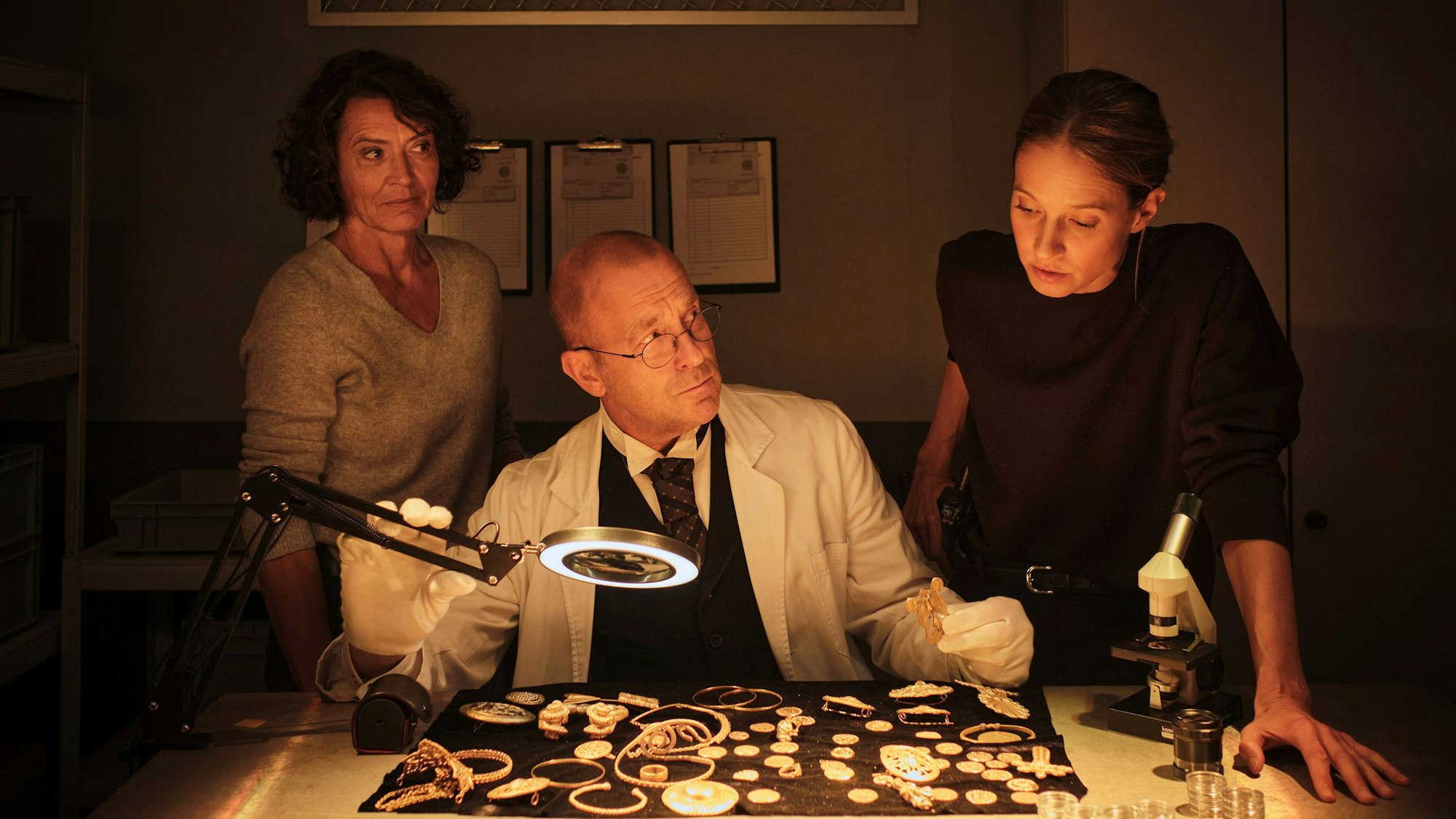 Johanna Stern beugt sich über den Goldschatz, der auf einer samtenen Decke liegt. Der Kurator Albert Dürr sitzt vor dem Schatz und beäugt Stern kritisch. Auch Odenthal schaut skeptisch zu ihrer Kollegin.