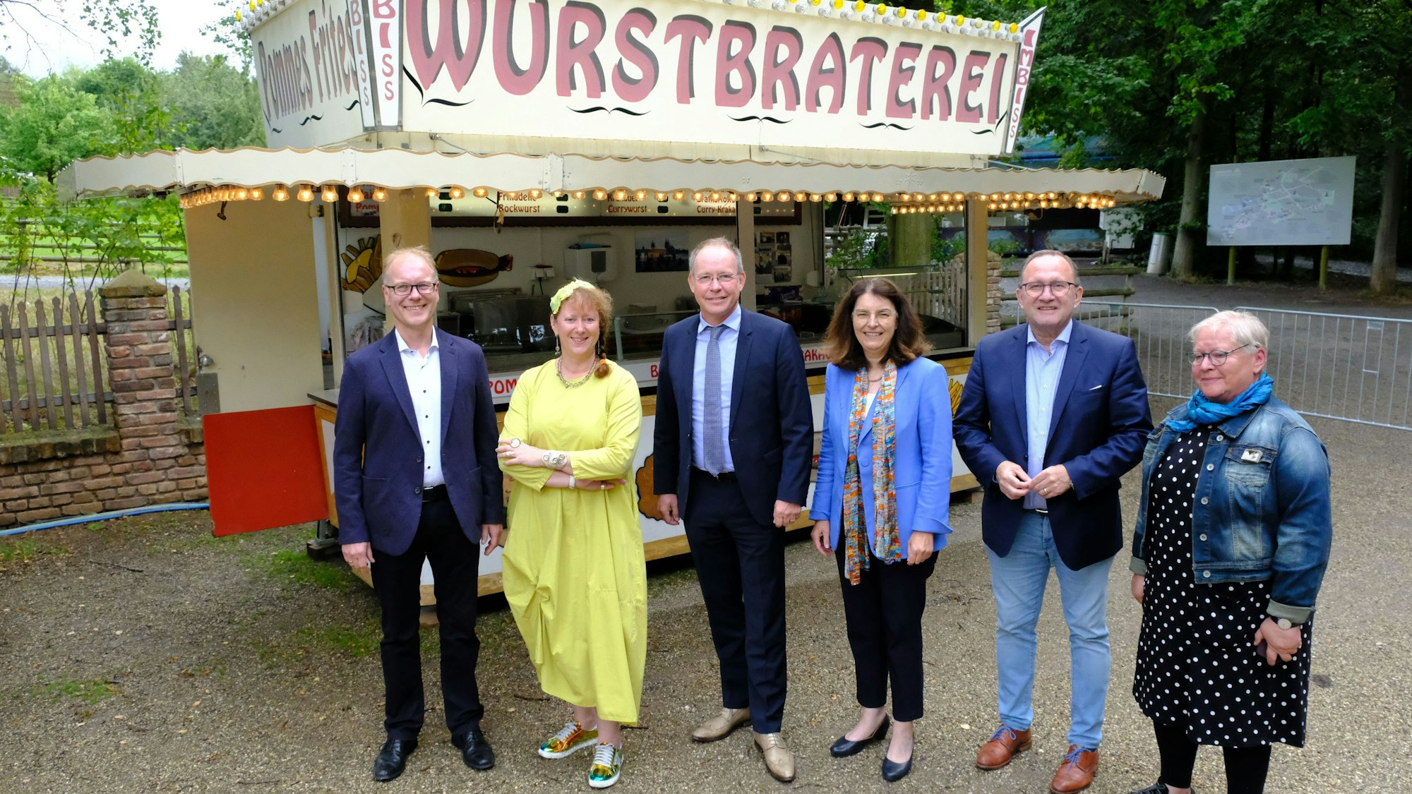 Museumsdirektor Dr. Carsten Vorwig, Staatssekretärin Andrea Milz, LVR-Landesrat Rainer Limbach (v.l.) und weitere Vertreter des Fördervereins des Freilichtmuseums Kommern stehen vor einer Imbissbude.