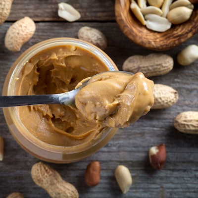 Ein Löffel Erdnussbutter liegt auf einem Glas Erdnussbutter. Um das Glas herum liegen Erdnüsse auf einem hölzernem Untergrund.