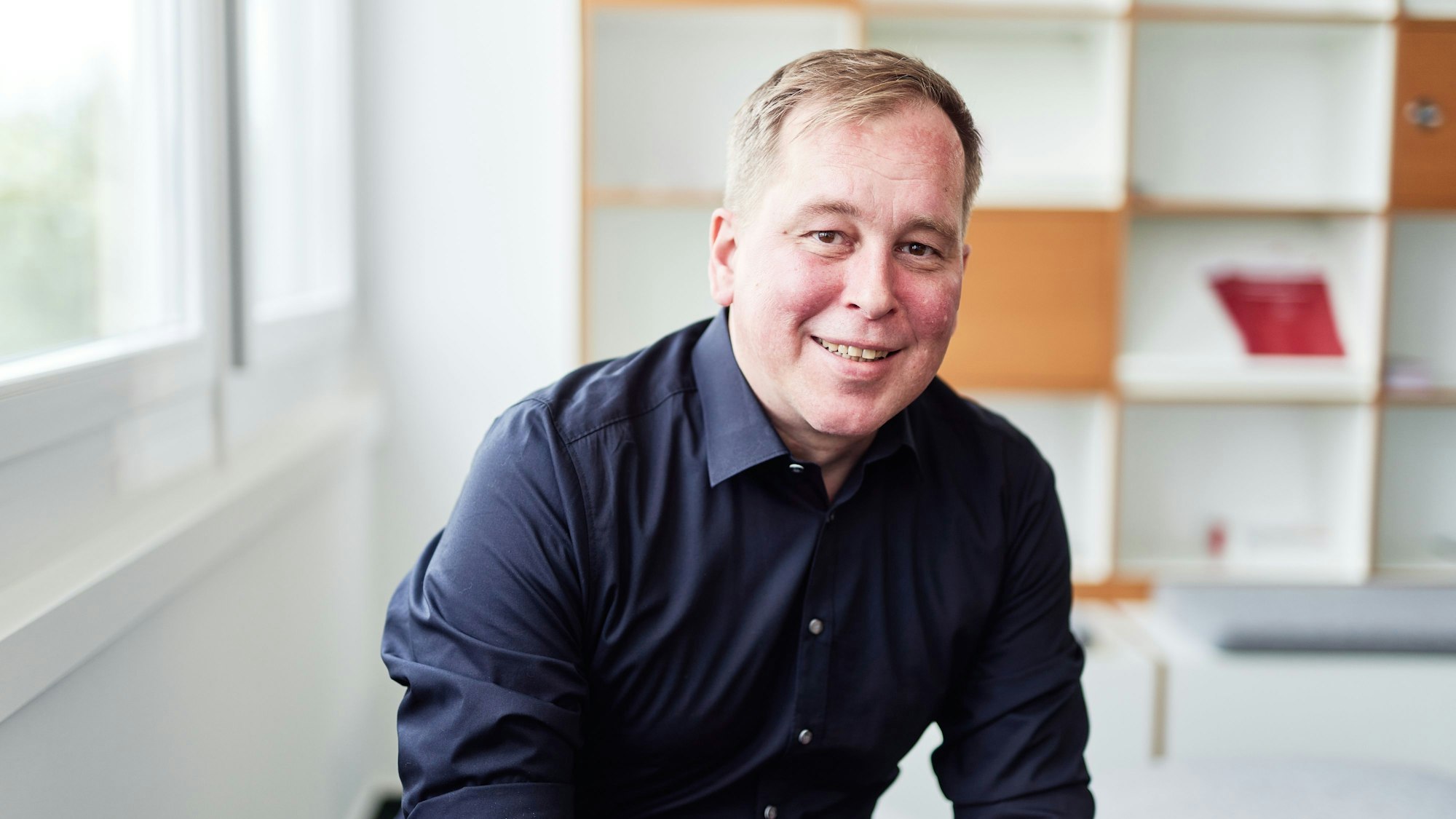 Florian Beer, Lehrer und pädagogischer Mitarbeiter von „Sabra" ist im Halbporträt zu sehen. Er sitzt in einem hellen Raum, trägt ein dunkles Hemd und lächelt in die Kamera. Hinter ihm steht ein weißes Bücherregal.