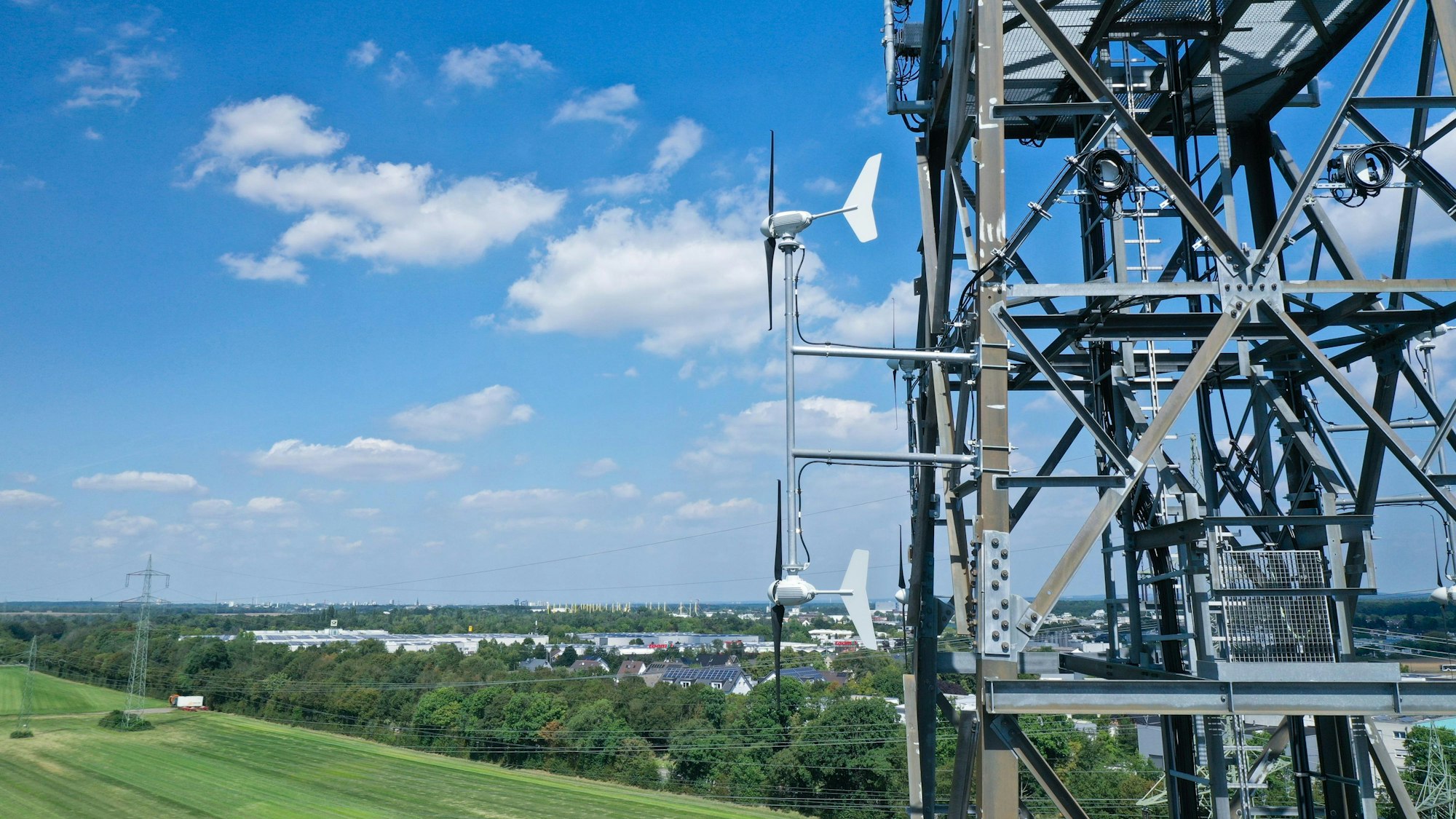 An einem Mobilfunkmast sind kleine Wind-Turbinen montiert. In der Ferne sieht man ein Gewerbegebiet, unten grüne Wiese.