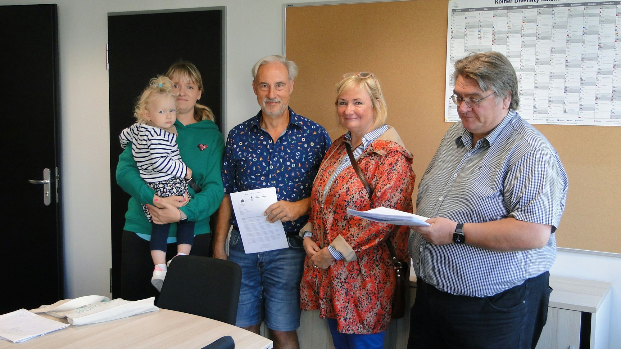 Eine Frau, mit einem Kind auf dem Arm, steht neben zwei Männern und einer Frau. Der Mann ganz rechts hält ein Papier in der Hand.