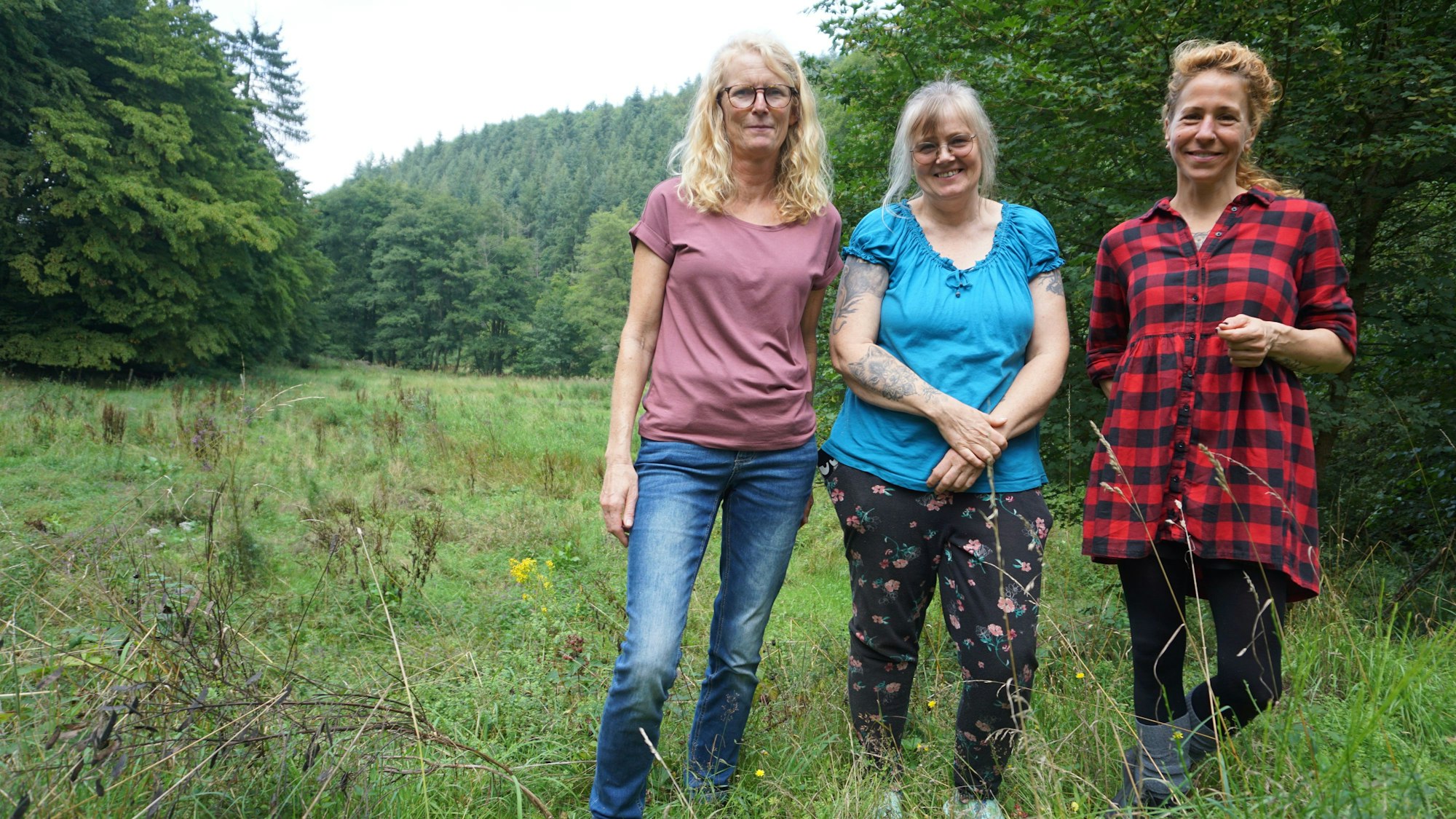 Drei Frauen stehen in Freizeitkleidung auf einer Wiese. Im Hintergrund ist Wald zu erkennen.