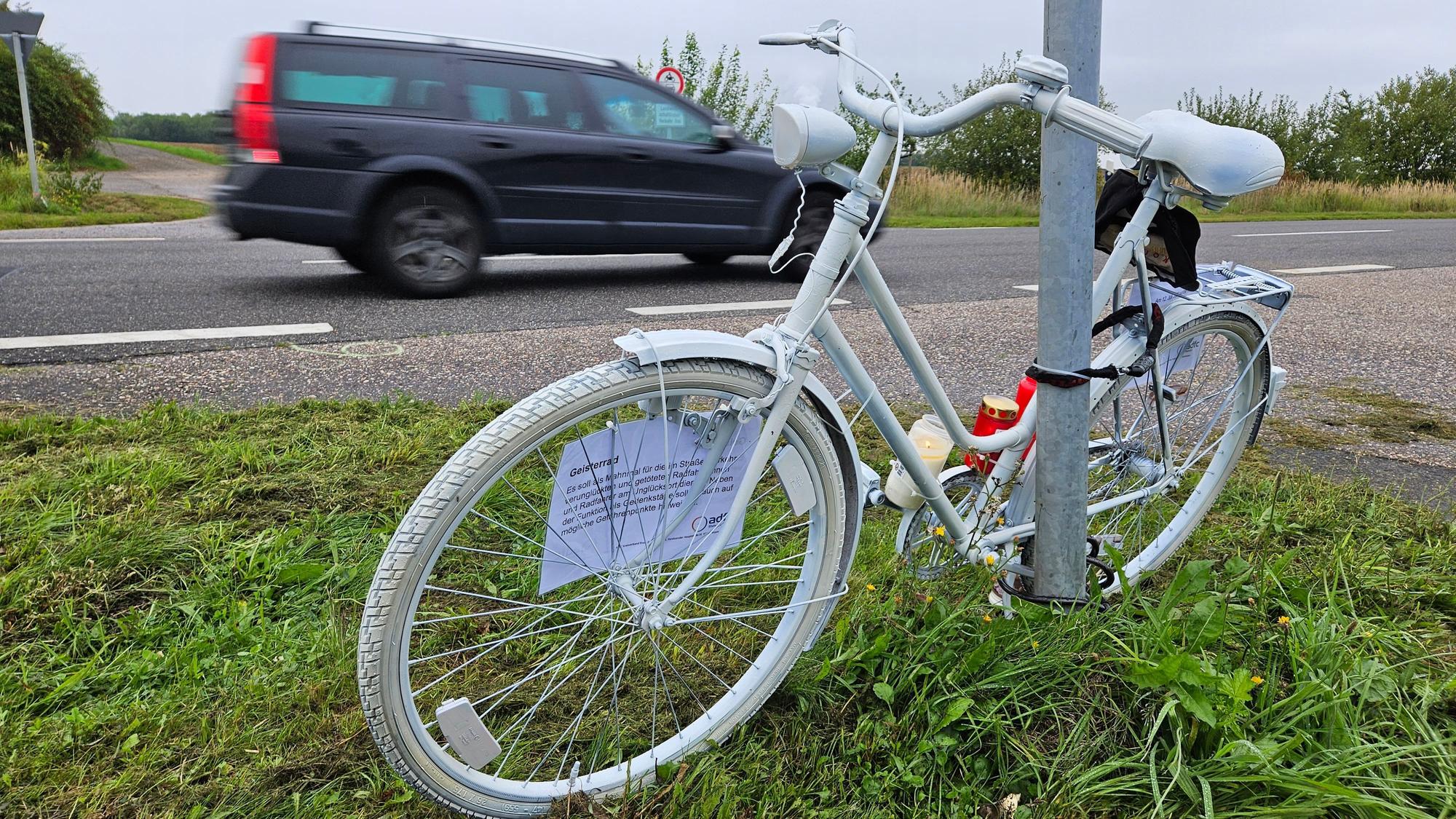 Ein komplett weiß angemaltes Fahrrad ist an einem Mast neben einer Straße angekettet, ein Auto fährt vorbei.
