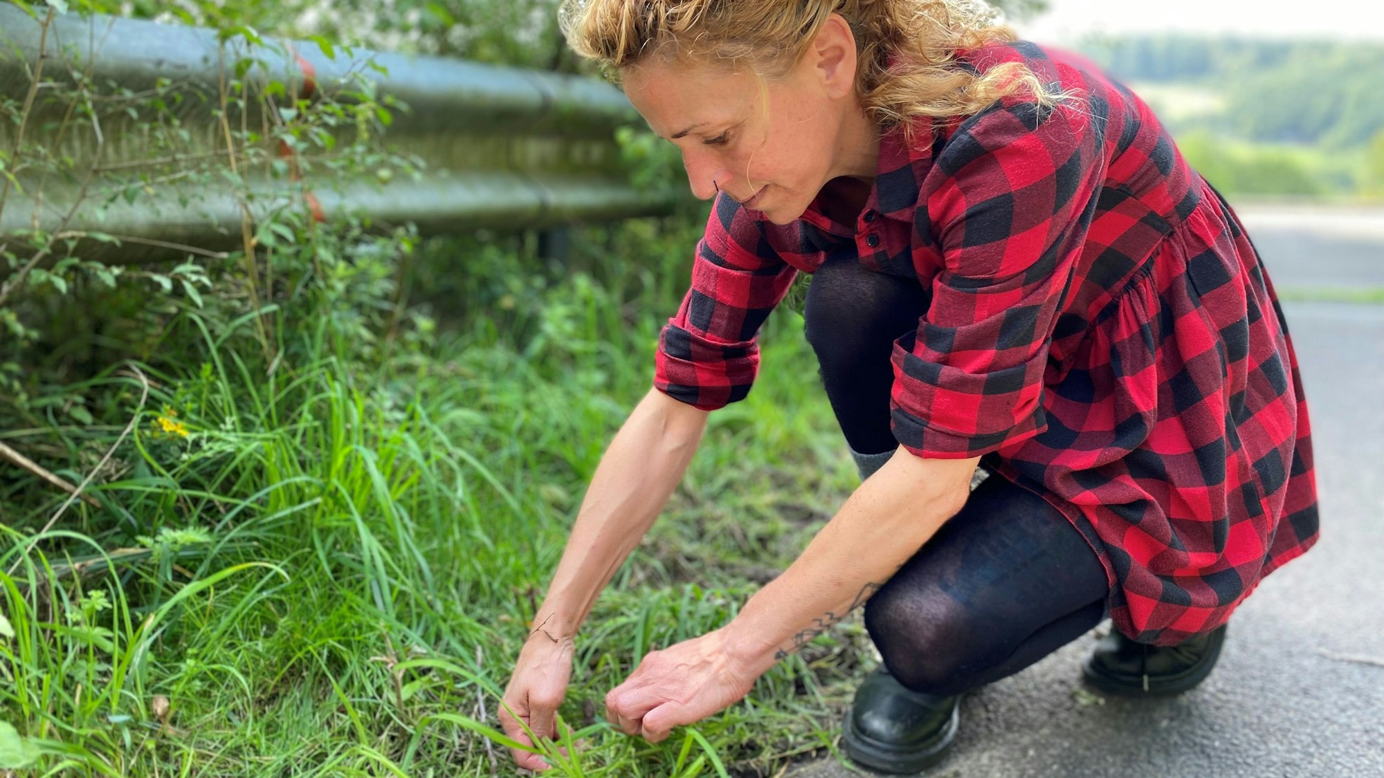 Eine Frau mit blonden Haaren kniet am Straßenrand und hält ihre Hände in das Gras. Sie trägt eine schwarze Hose und ein rot-kariertes Hemd.