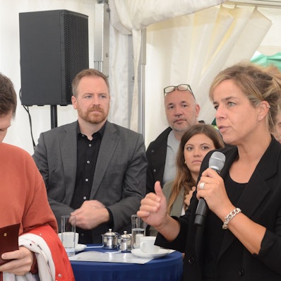 Die Grüne NRW-Wirtschaftsministerin spricht in einem Zelt. Sie hat ein Mikrofon in der Hand. Ein Mann im roten Pullover macht sich Notizen.
