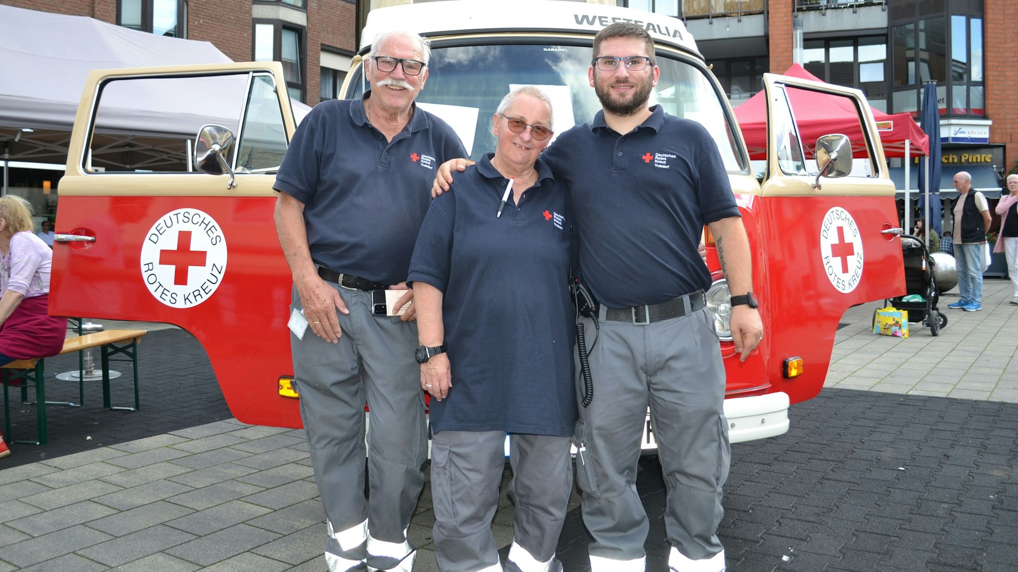 Drei Männer in den Uniformen des DRK stehen vor einem roten VW-Bulli-Oldtimer.