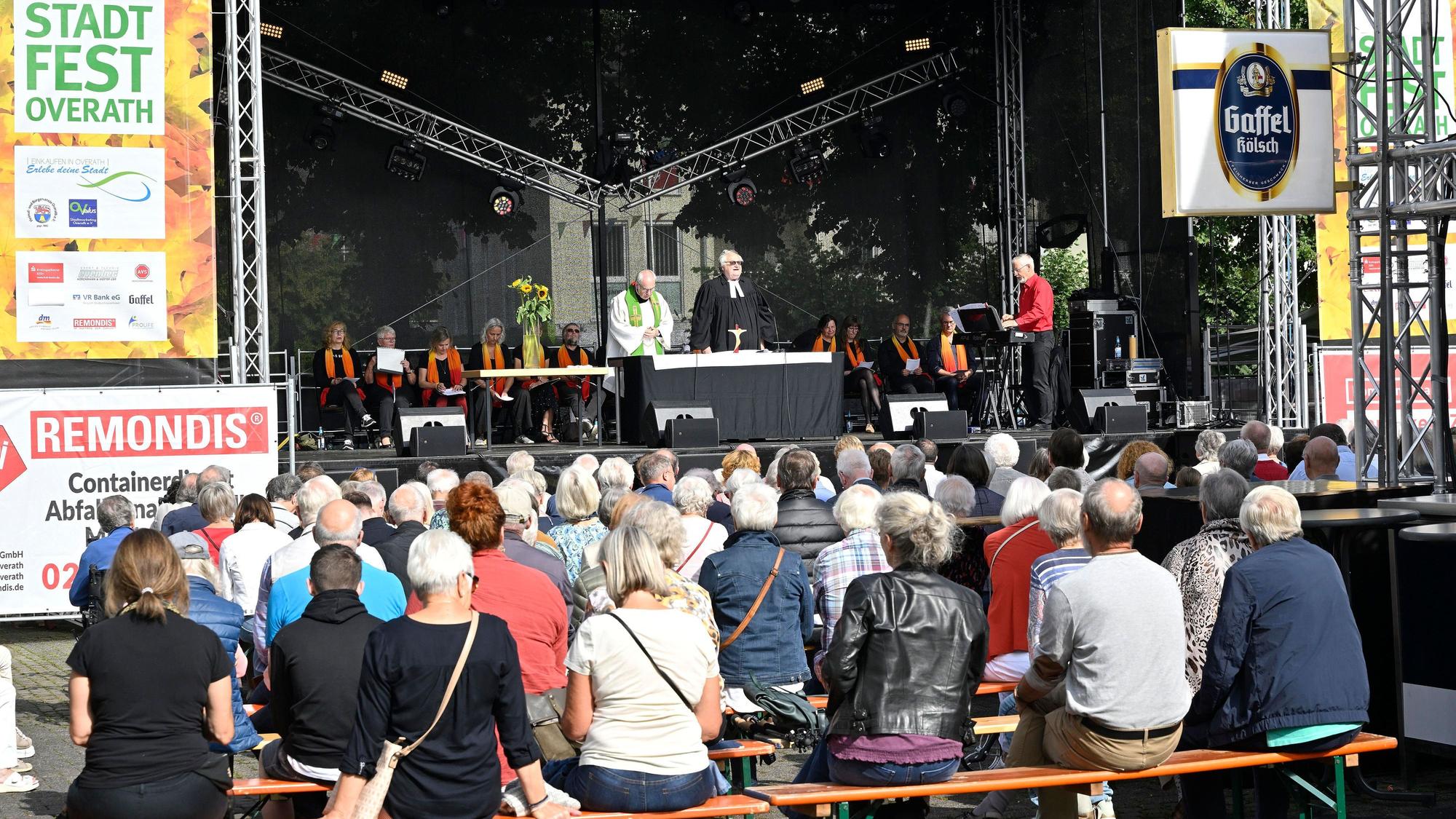 Menschen sitzen auf Bänken und hören den beiden Priestern zu, die auf der Bühne beim Overather Stadtfest einen ökumenischen Gottesdienst zelebrieren.