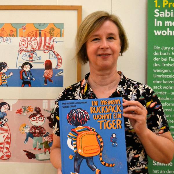 Eine blonde Frau hält das blaue Bilderbuch. Neben ihr hängen zwei Illustrationen aus ihrem Buch, die den katzenähnlichen Tiger mit Kindern zeigen.