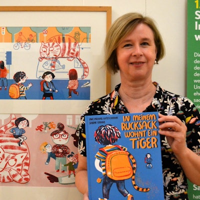 Eine blonde Frau hält das blaue Bilderbuch. Neben ihr hängen zwei Illustrationen aus ihrem Buch, die den katzenähnlichen Tiger mit Kindern zeigen.