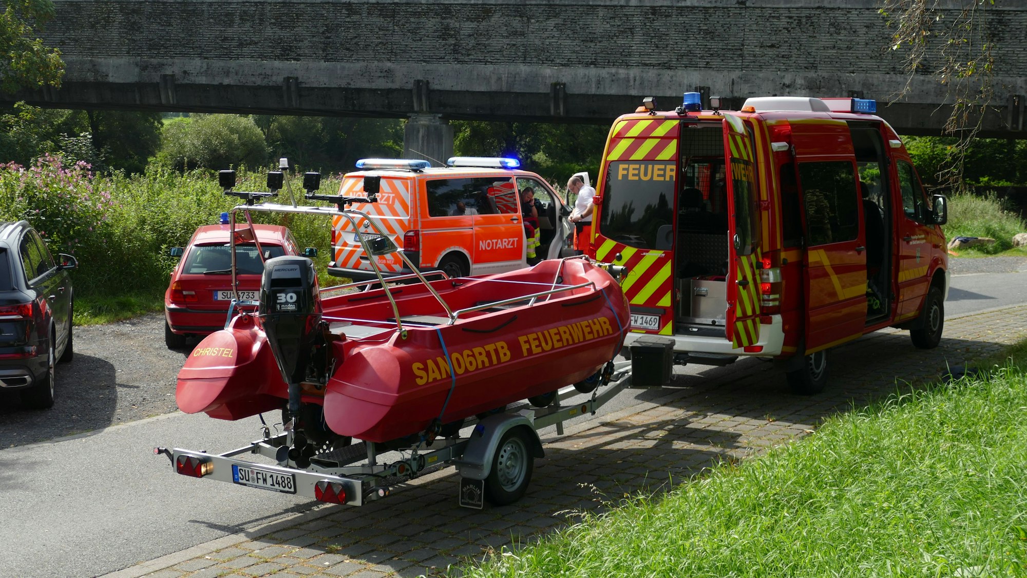 Auf einer Straßen an der Sieg stehen Fahrzeuge des Rettungsdienstes und der Feuerwehr, diese hat ein Schlauchboot auf einem Anhänger.