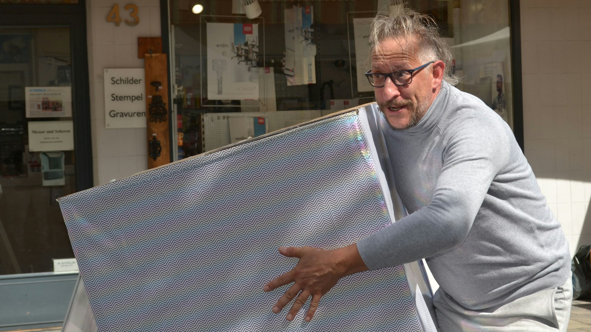 Ein Mann in grauem Pullover und grauer Hose hält erwartungsvoll eine große graue Kiste.