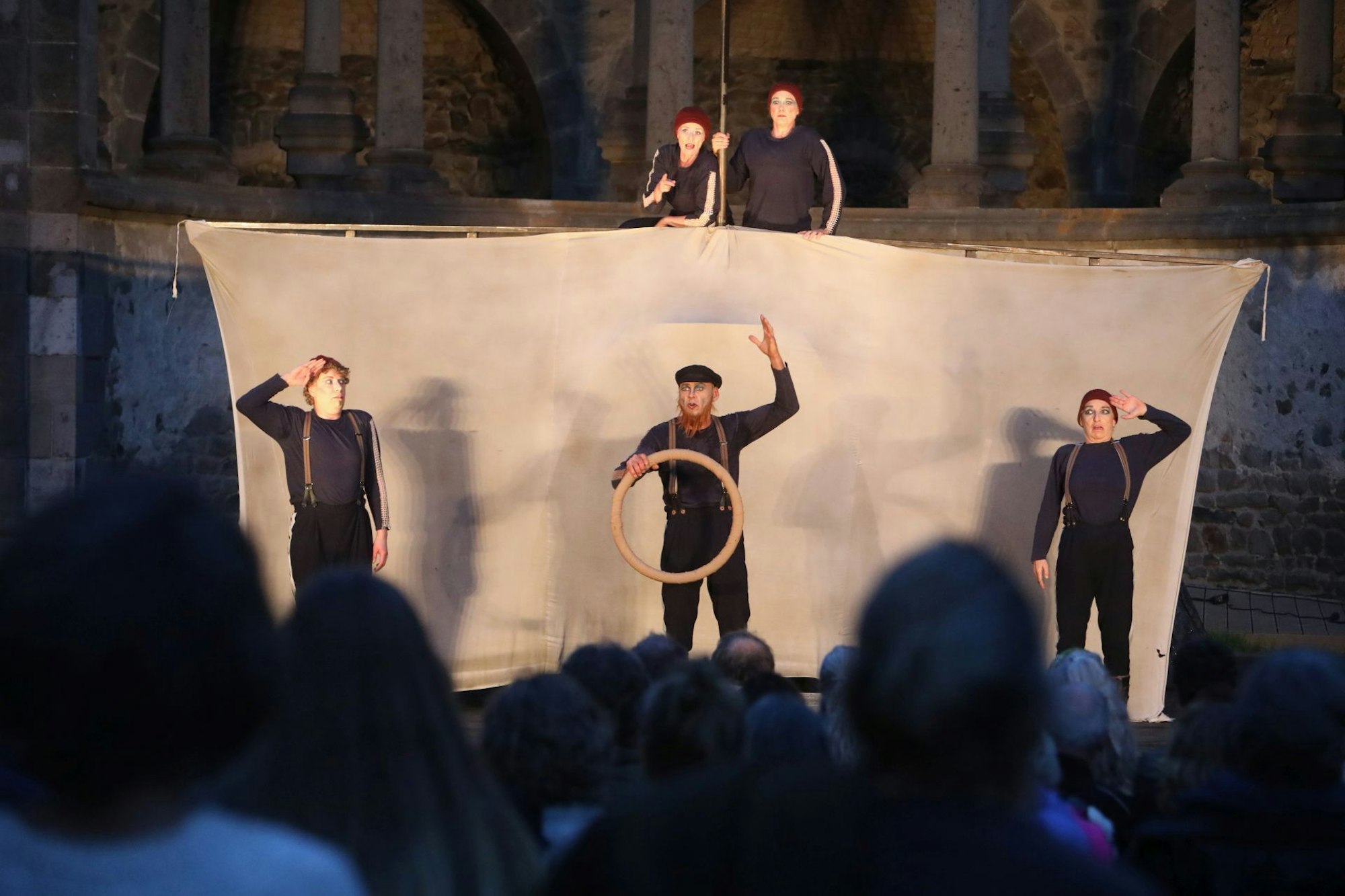Drei Schauspieler vor einem Segel, zwei obenauf — im Hintergrund ist die Ruine des Abteichors von Kloster Heisterbach zu erkennen.