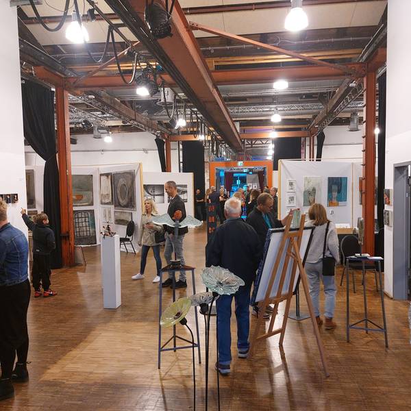 Eine große Ausstellungshalle mit Holzboden und einem Dach aus Stahlträgern. Skulpturen und Staffeleien stehen im Raum, an den Wänden hängen Bilder. Viele Menschen betrachten die Kunstwerke.
