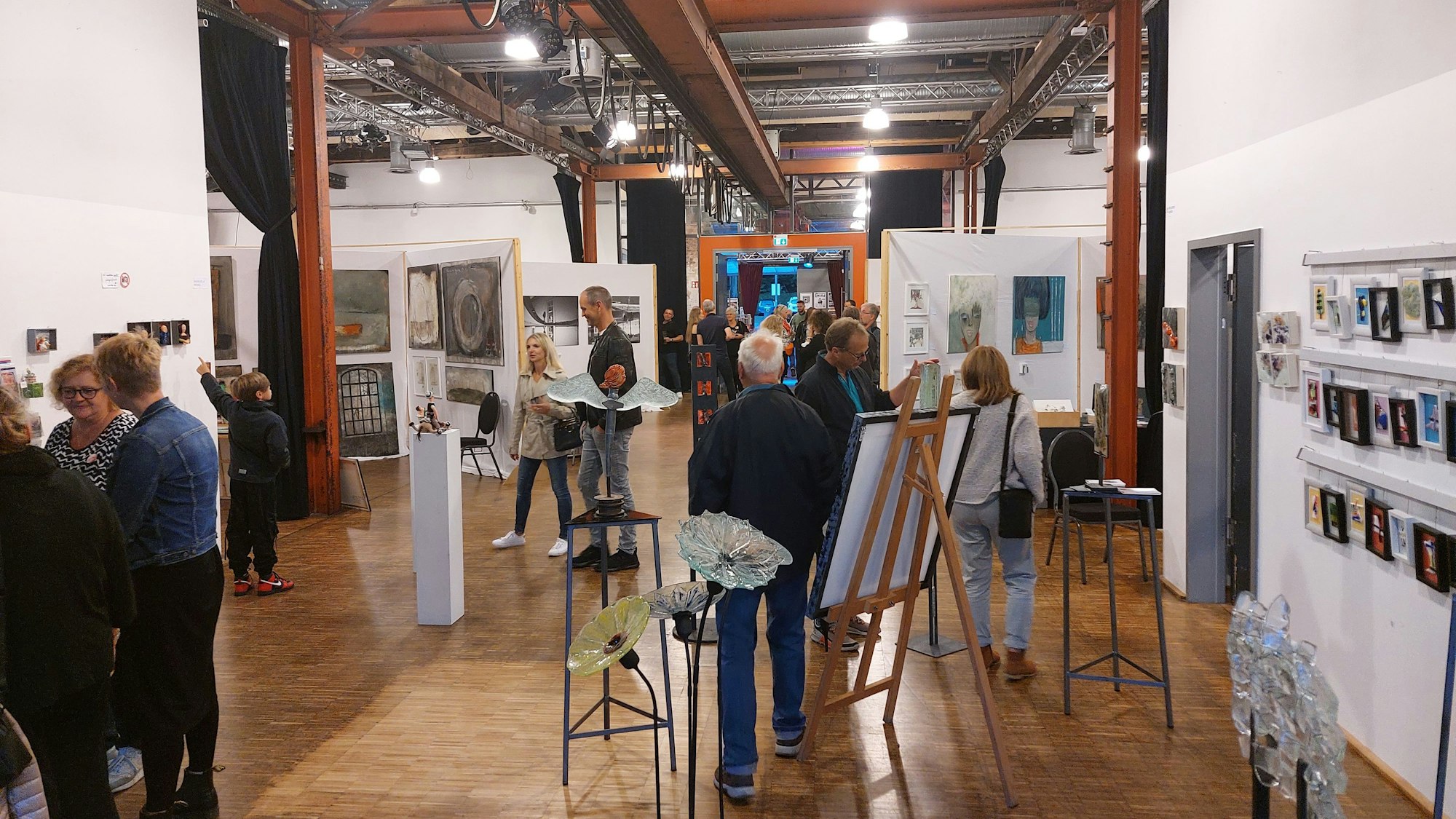 Eine große Ausstellungshalle mit Holzboden und einem Dach aus Stahlträgern. Skulpturen und Staffeleien stehen im Raum, an den Wänden hängen Bilder. Viele Menschen betrachten die Kunstwerke.