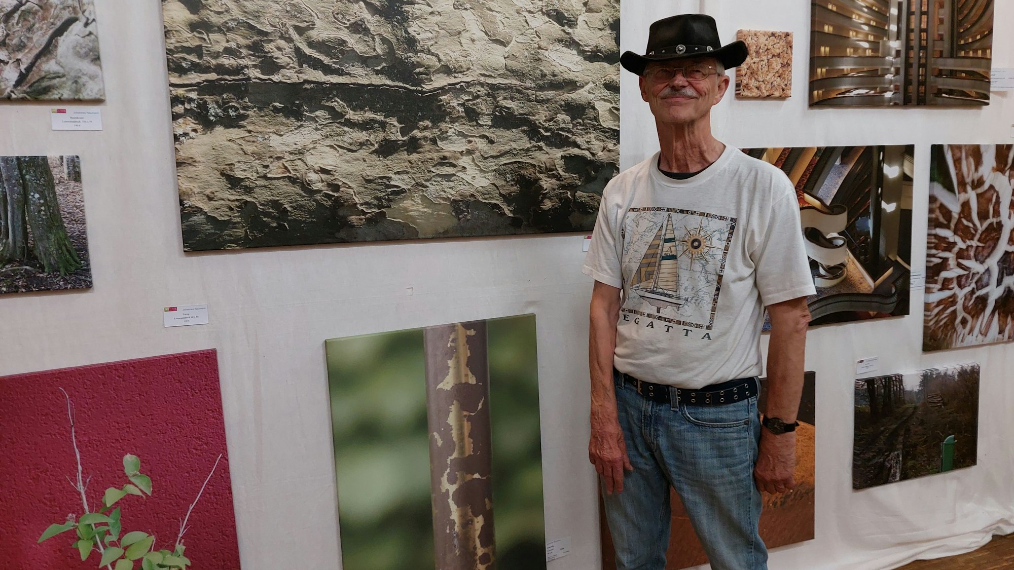 Ein älterer Mann mit Cowboyhut vor großen Fotoggrafien, die Details von Wänden, Pflanzen oder Rohren zeigen.