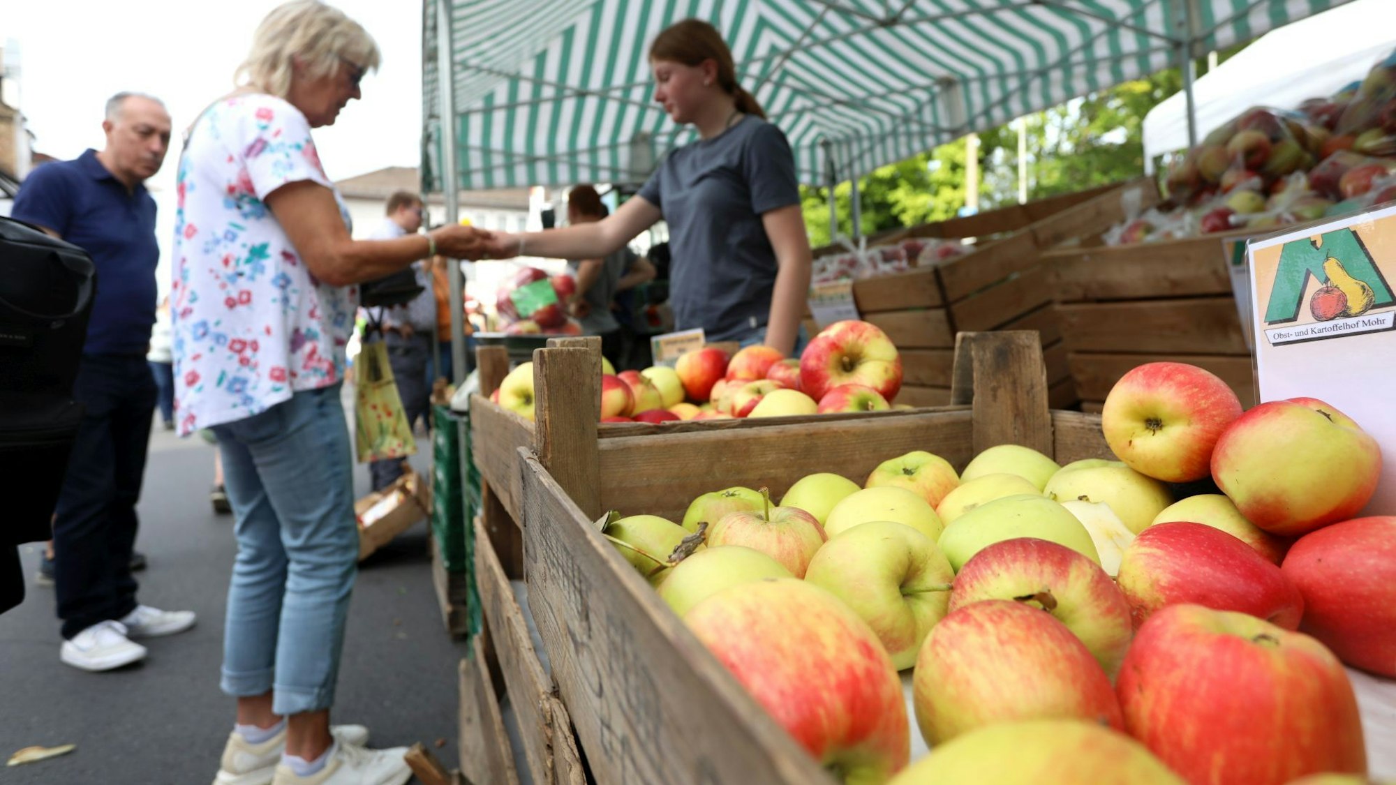Rotbackige Äpfel liegen in Holzkisten, eine Frau gibt einer Verkäuferin Geld.