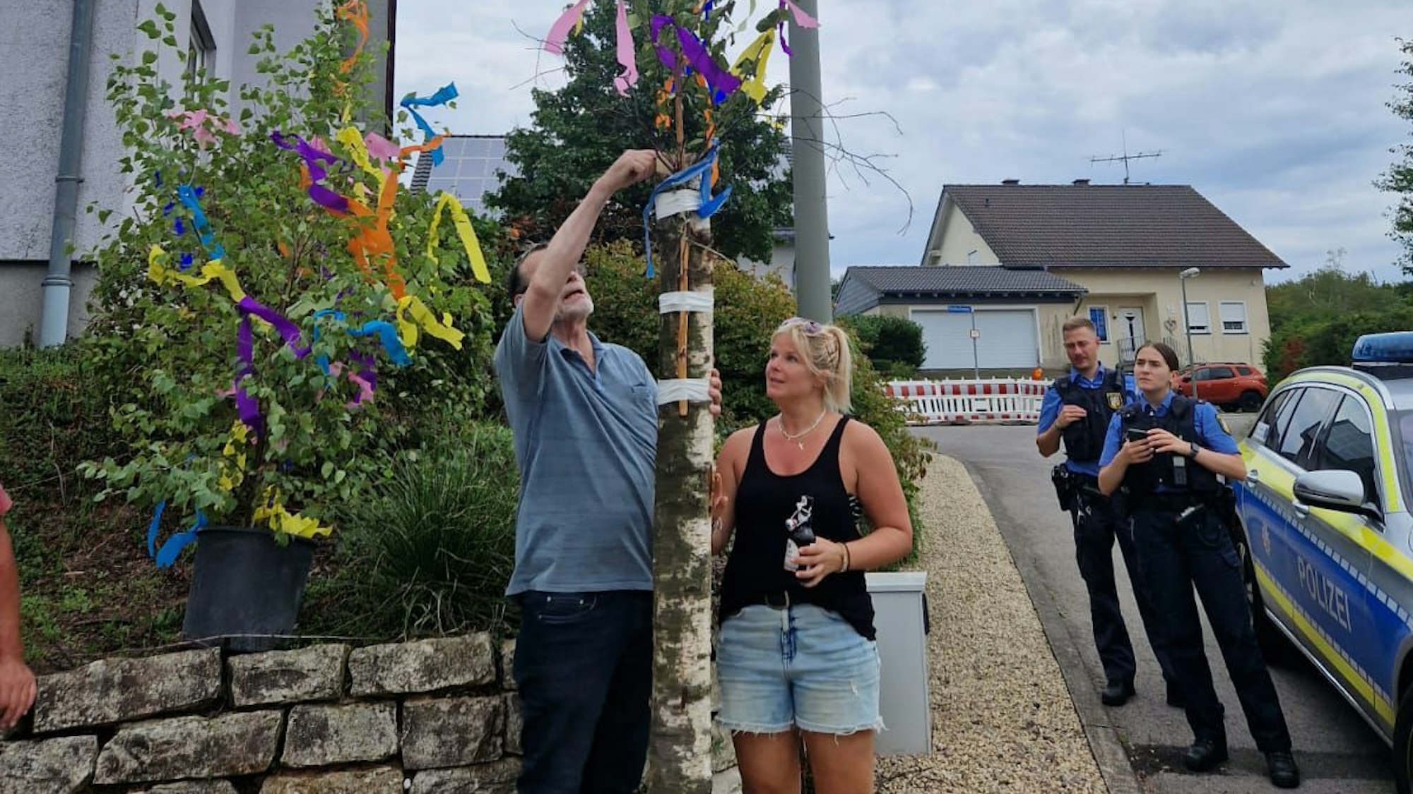 Ein Mann und eine Frau stehen links und rechts von einem Birkenstamm ohne Krone. Eine Polizistin und Polizist stehen daneben.
