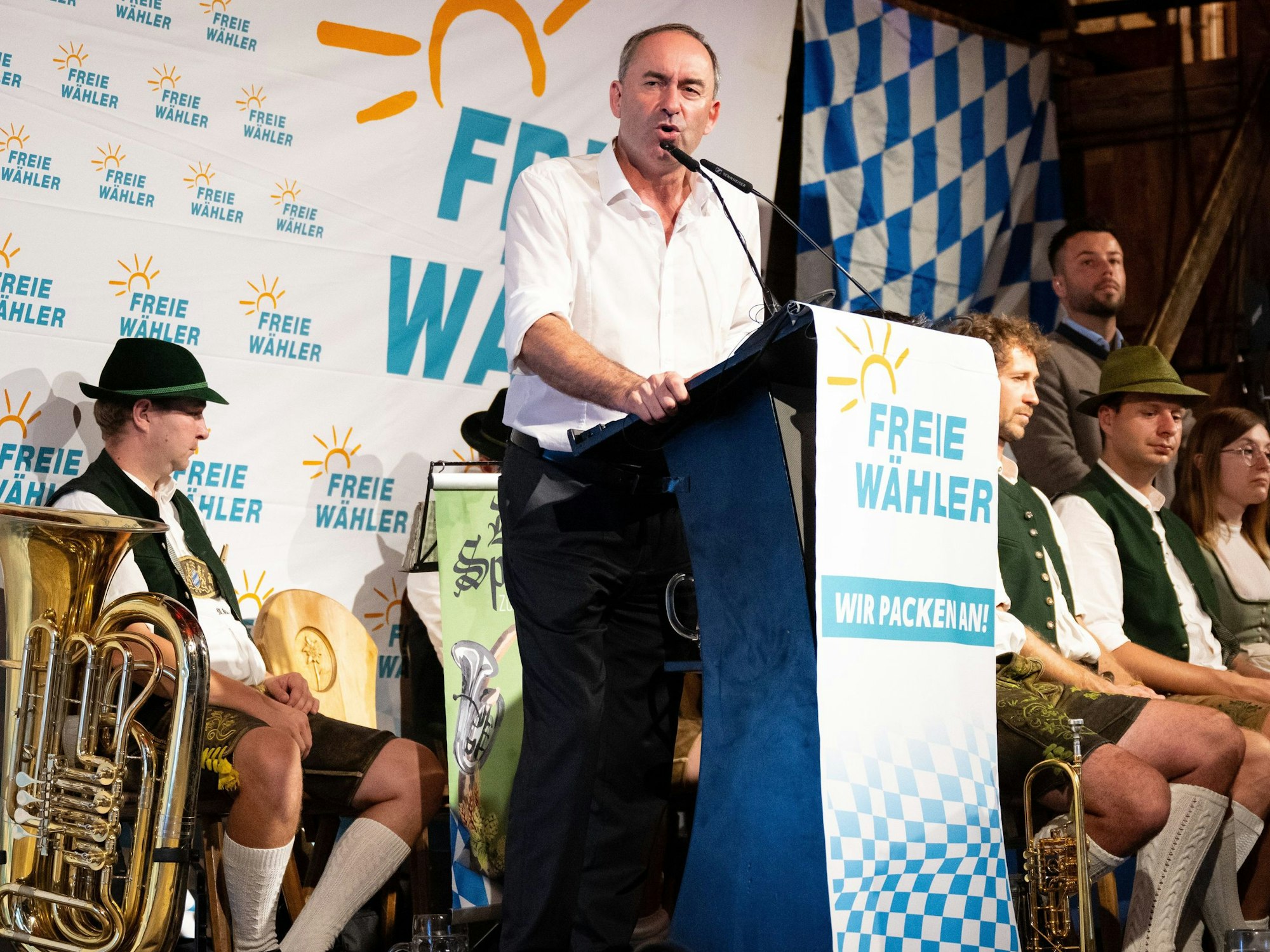 Hubert Aiwanger, Bundesvorsitzender der Freien Wähler und stellvertretender Ministerpräsident von Bayern, spricht bei einem Volksfest in Niederbayern von einer Tribüne. Hinter und neben ihm sitzen Musiker in Trachtenanzügen mit Blasinstrumenten. Die Wand im Bildhintergrund ist mehrfach mit einem Sonnenemblem und dem Schriftzug "Freie Wähler" bedruckt.