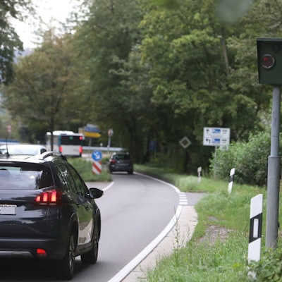 Eine gebogene Straße, auf der ein Auto fährt. Am Straßenrand steht ein grüner Radarkasten, daneben ein gelbes Schild mit der Richtung Königswinter.