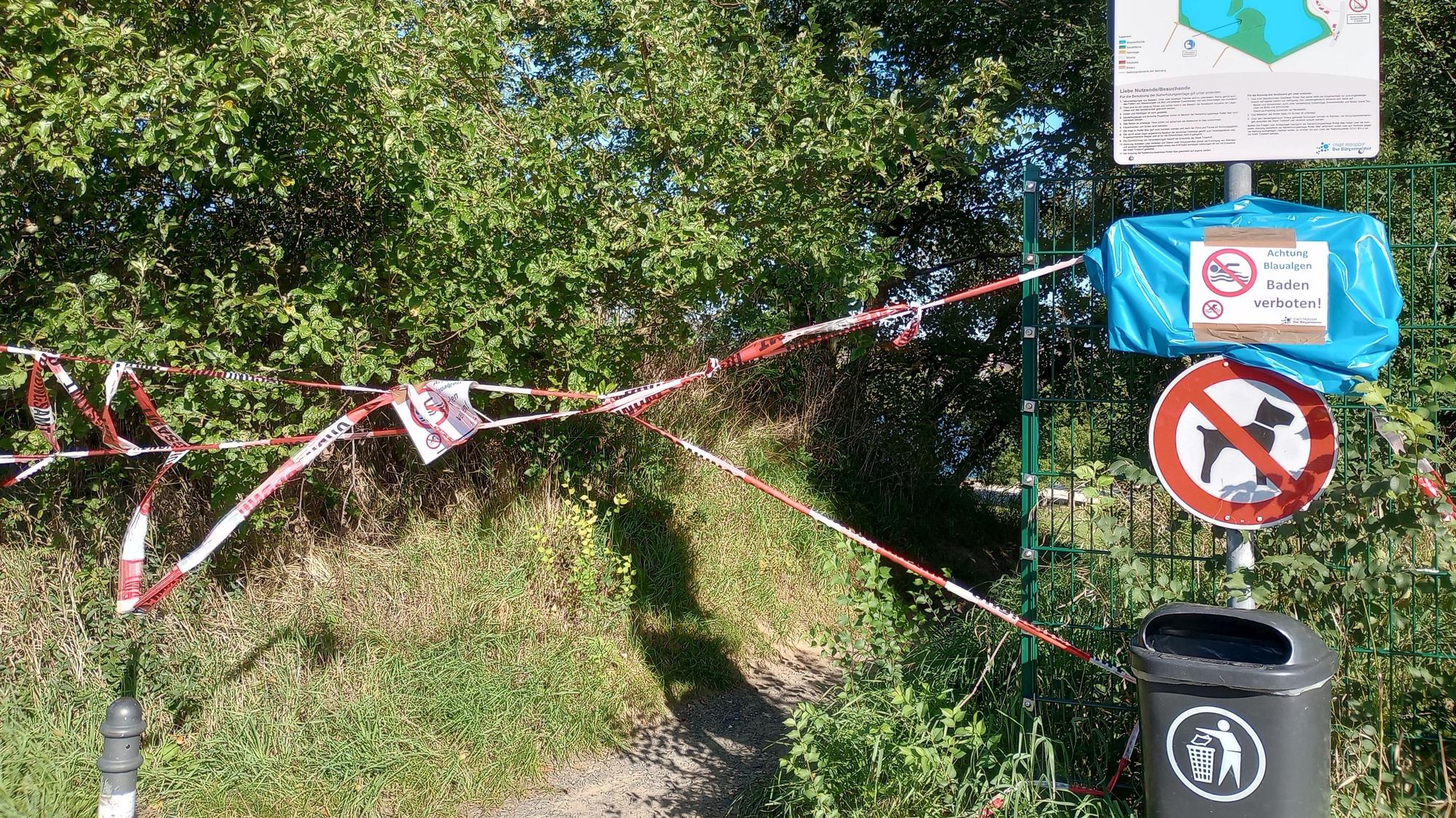 Rot-weißes Flatterband sperrt einen Fußweg ab.An einem Metallpfosten sind eine Karte des Sees, ein Warn- und Verbotsschild und ein Papierkorb befestigt. Auch Hunde sind hier nicht gestattet.