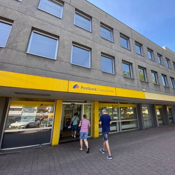 Drei Menschen betreten eine Filiale der Postbank in einem grauen Gebäude.