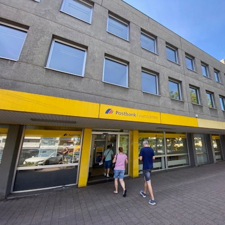 Drei Menschen betreten eine Filiale der Postbank in einem grauen Gebäude.