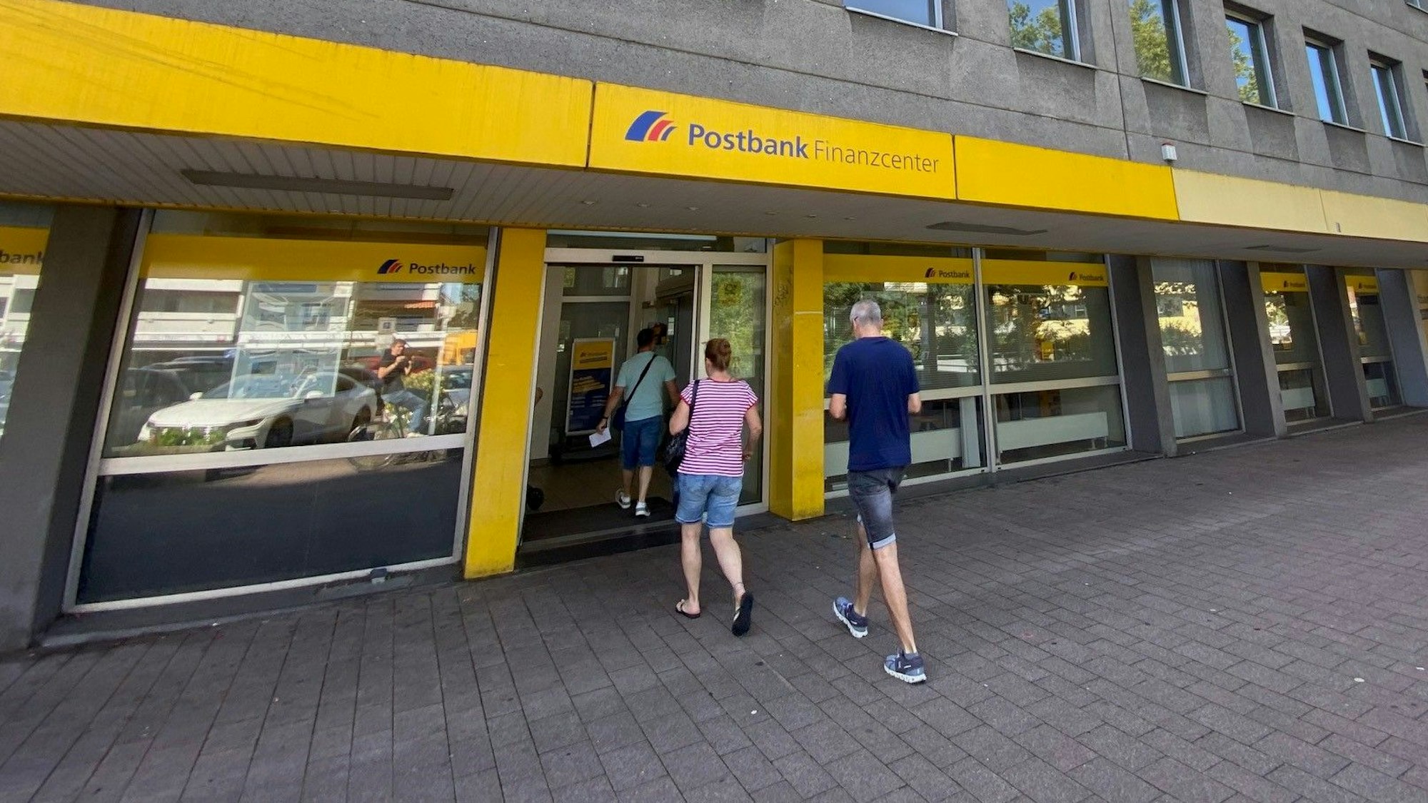 Drei Menschen betreten eine Filiale der Postbank in einem grauen Gebäude.