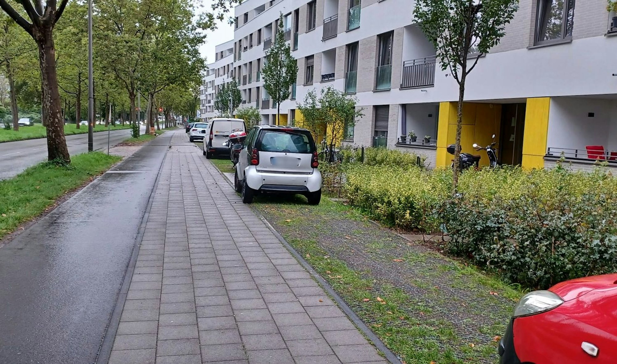 Vor einem Wohnblock parken Autos auf einer Schotterfläche neben Rad- und Fußweg.