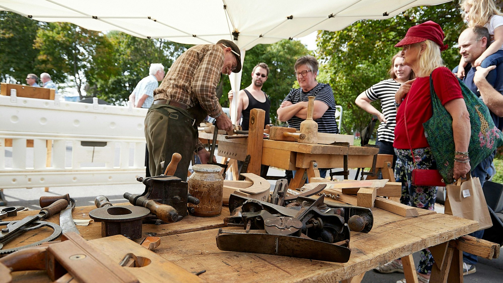 Im Vordergrund liegen Werkzeuge auf einem Tisch, im Hintergrund arbeitet ein Schreiner. Leute schauen zu.