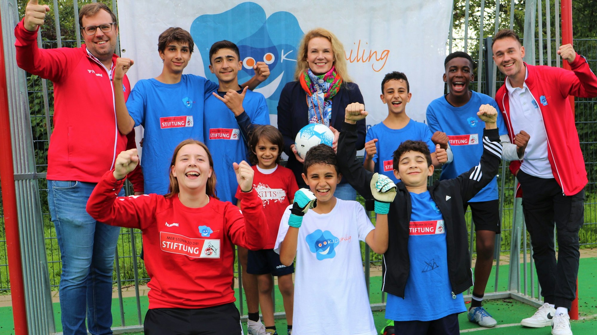 Junge Fußballer von dem Kölner Verein „Kidsmiling“ und Vereinsmitarbeiterinnen und -Mitarbeiter stehen vor einem Tor in Siegerposen.