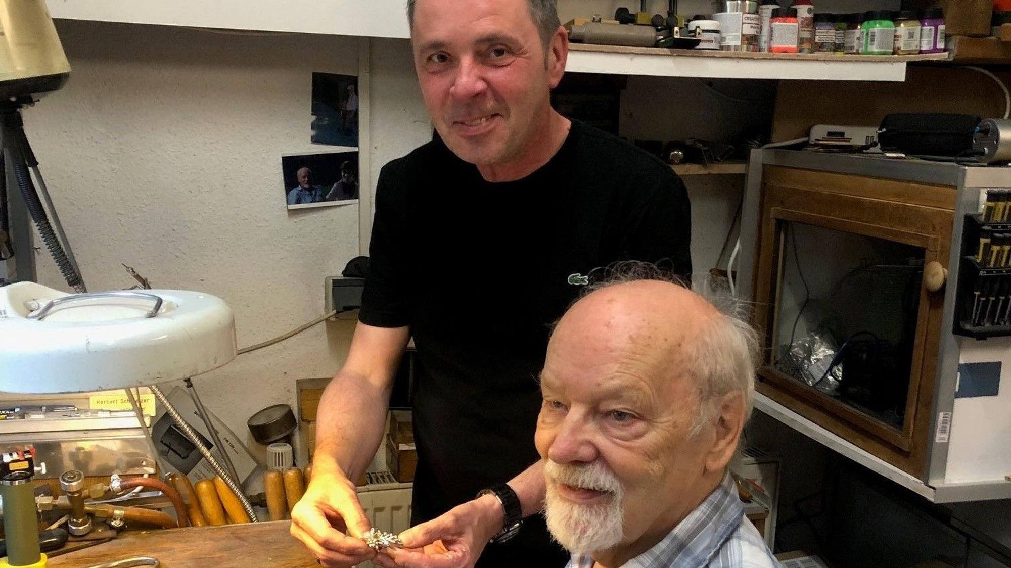 Friseurmeister Ralph Schwalbach zeigt Goldschmied Herbert Schmelzer eine Brosche, die er zugunsten von „wir helfen“ verkaufen möchte