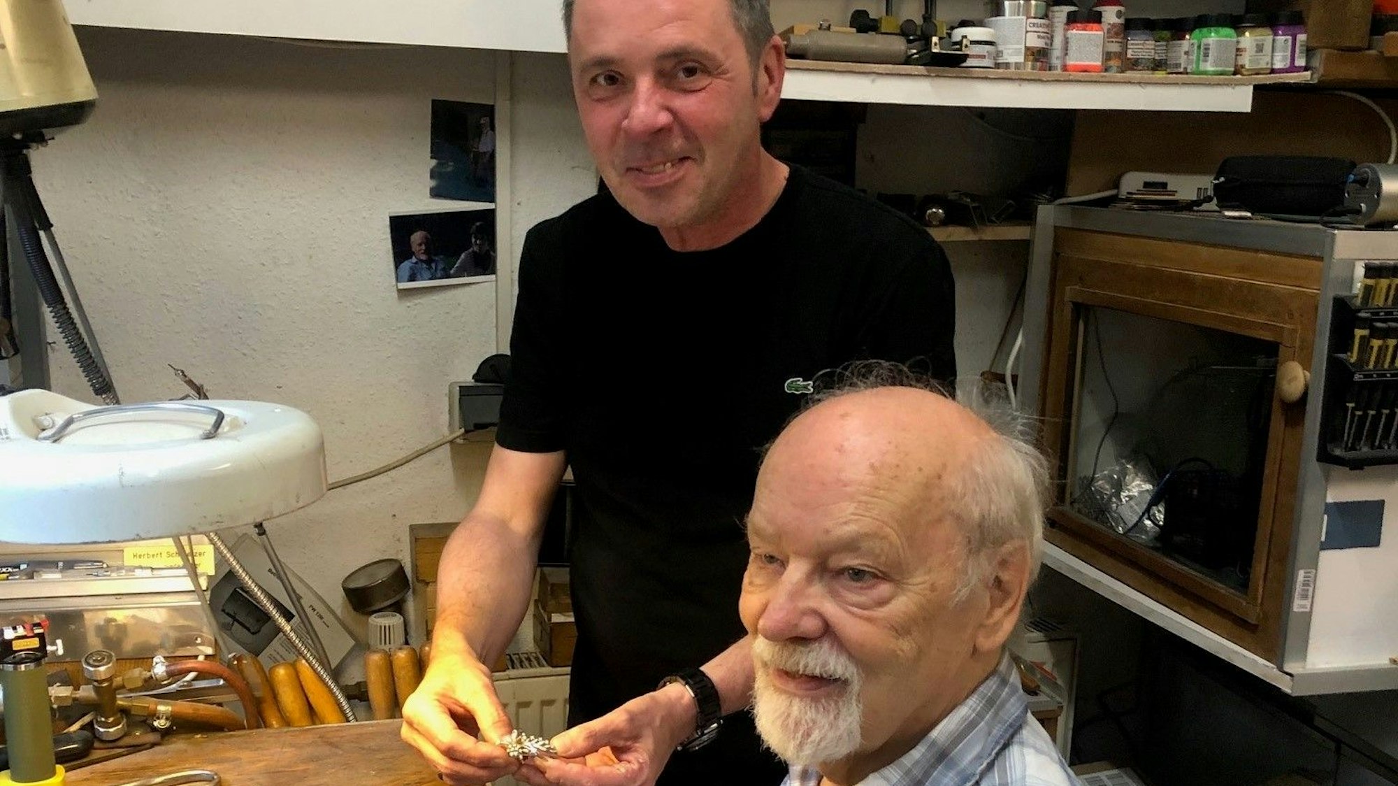 Friseurmeister Ralph Schwalbach zeigt Goldschmied Herbert Schmelzer eine Brosche, die er zugunsten von „wir helfen“ verkaufen möchte