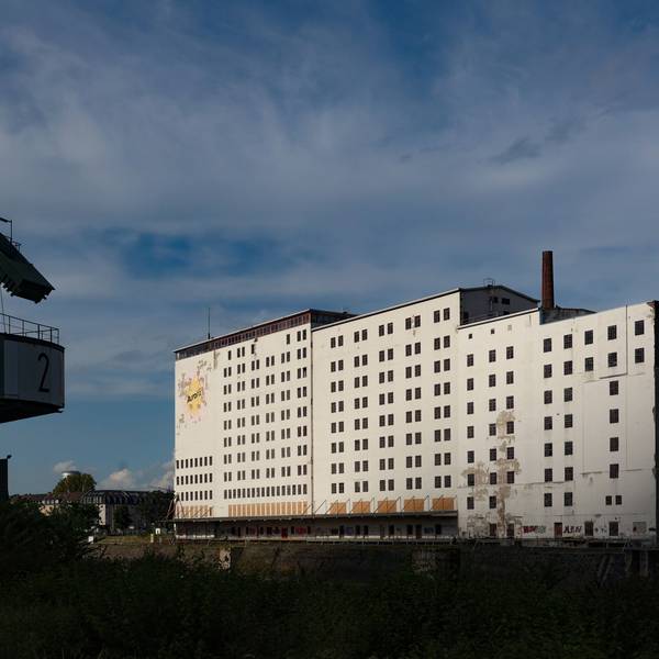 Ein großes, wuchtiges, weißes Gebäude steht am Deutzer Hafenbecken. Im Vordergrund ist ein ausgemusterter Hafenkran zu sehen.