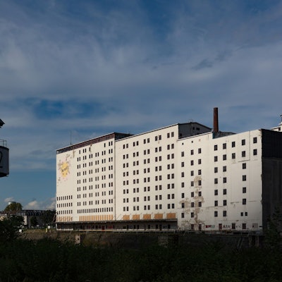 Ein großes, wuchtiges, weißes Gebäude steht am Deutzer Hafenbecken. Im Vordergrund ist ein ausgemusterter Hafenkran zu sehen.