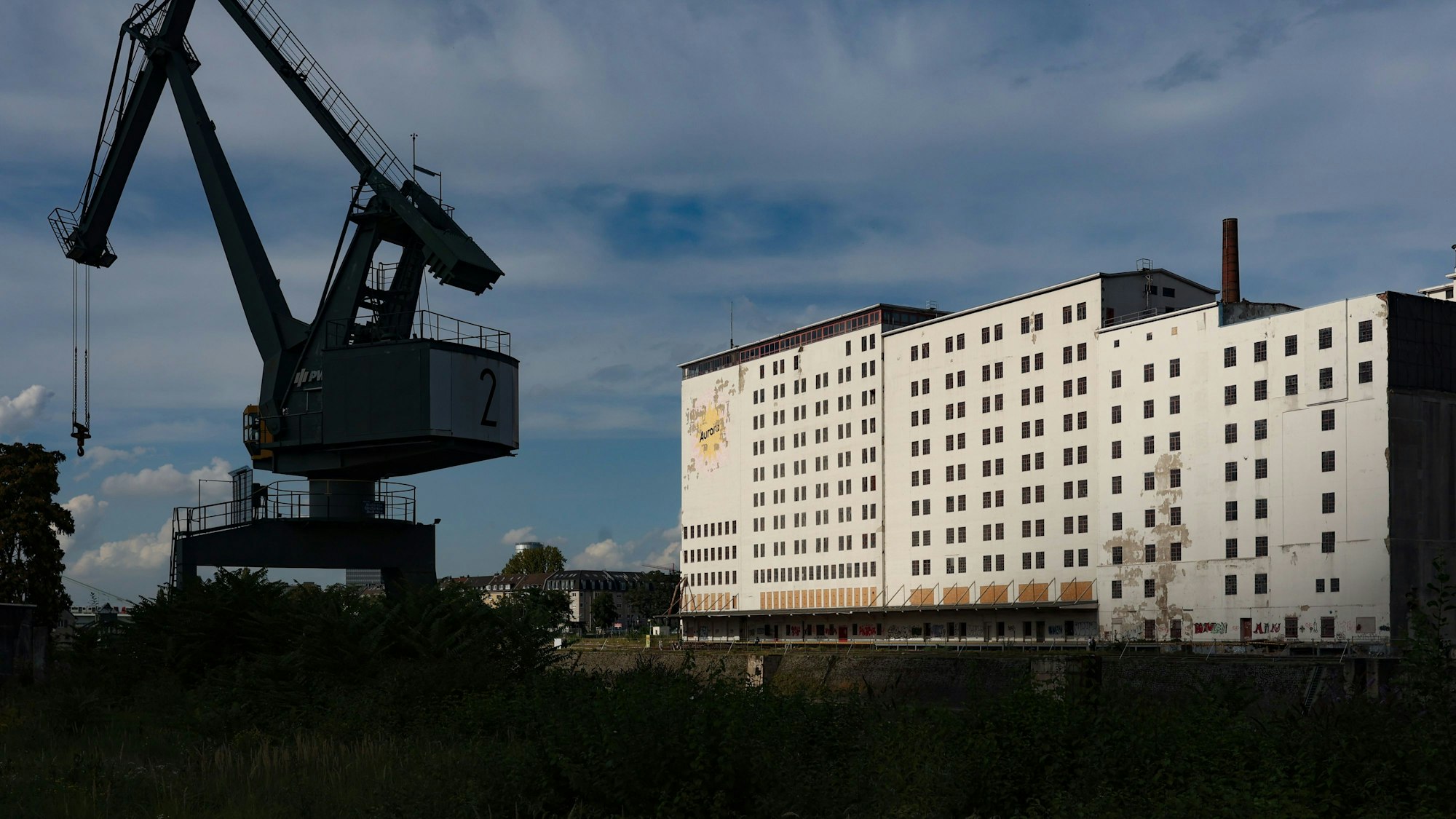 Ein großes, wuchtiges, weißes Gebäude steht am Deutzer Hafenbecken. Im Vordergrund ist ein ausgemusterter Hafenkran zu sehen.
