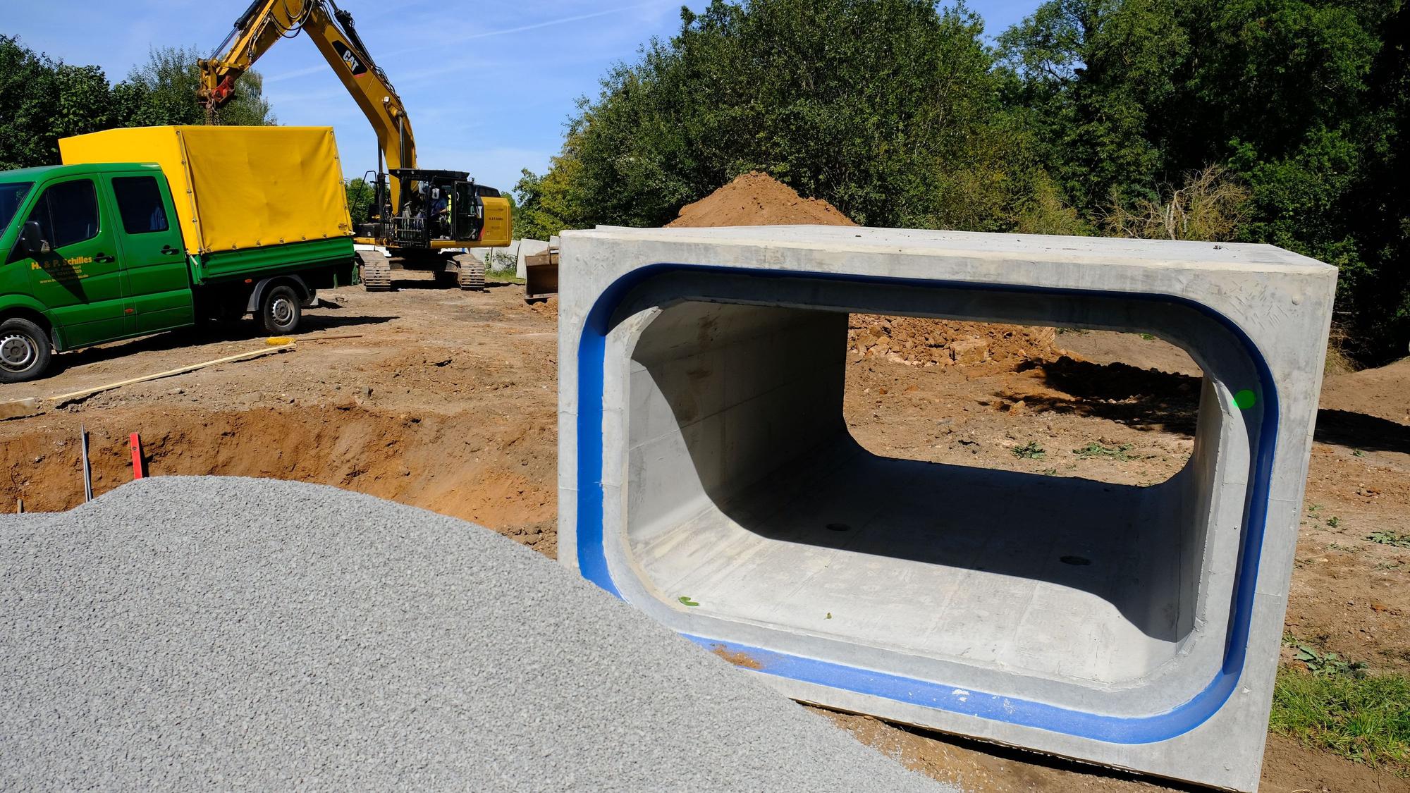 Ein kastenförmiges Rohr liegt auf einer Baustelle in Satzvey. Ein Haufen Splitt sowie im Hintergrund ein Lkw und ein Bagger sind zu sehen.