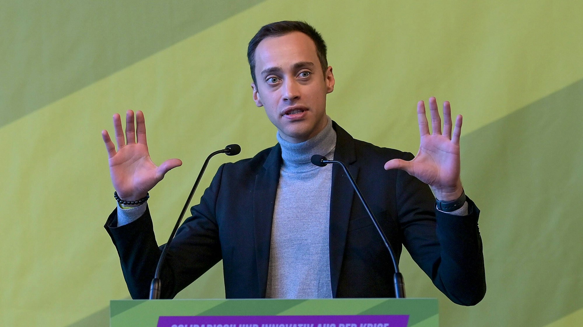 Tim Achtermeyer, Landesvorsitzender der Grüne NRW, ist an einem Rednerpult zu sehen. Er trägt einen grauen Rollkragenpullover und ein dunkles Jackett.