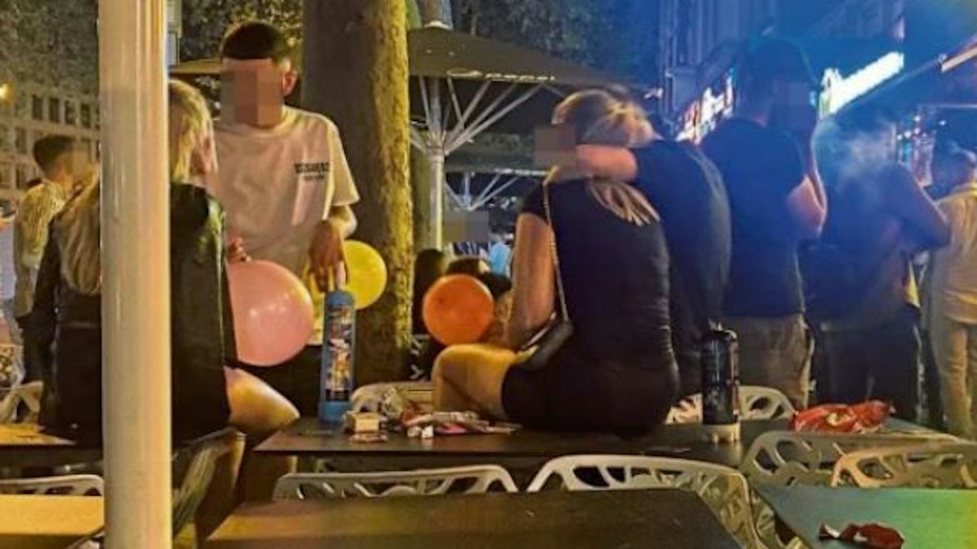 Junge Männer und Frauen sitzen mit Luftballons voller Lachgas auf Tischen an den Kölner Ringen.