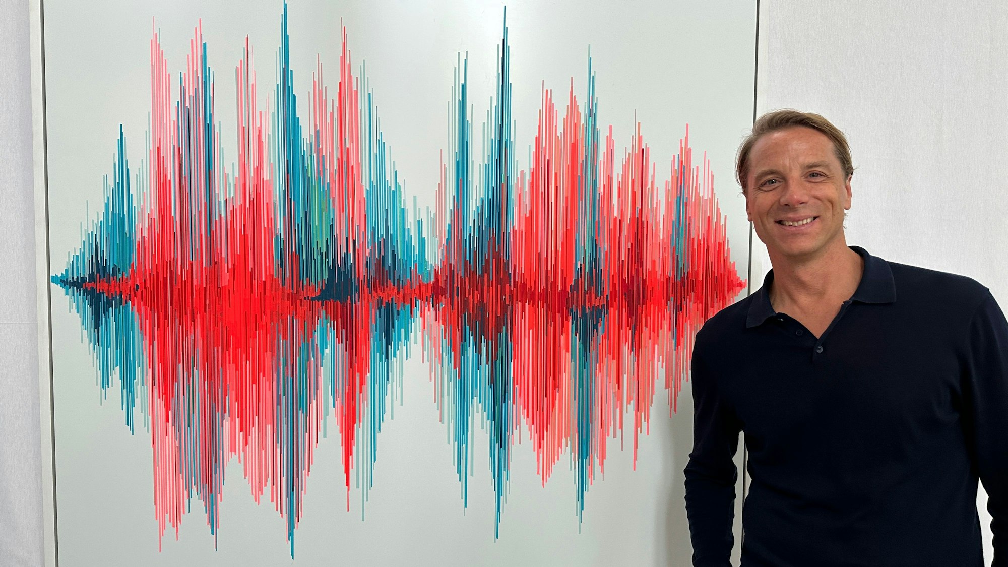 Ein Mann mit braunem Haar und schwarzem Pullover steht neben einem Bild.