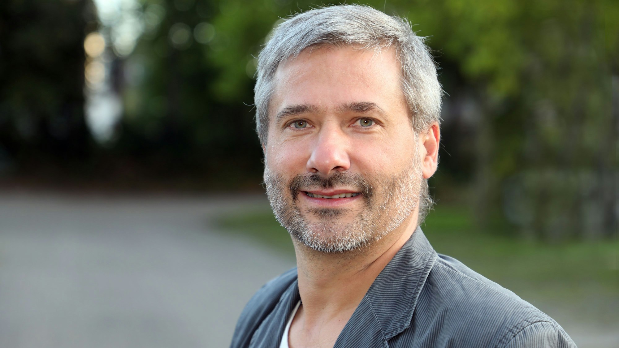 Ein Mann mit Bart schaut in die Kamera, im Hintergrund sind unscharf Bäume und eine Straße zu sehen.