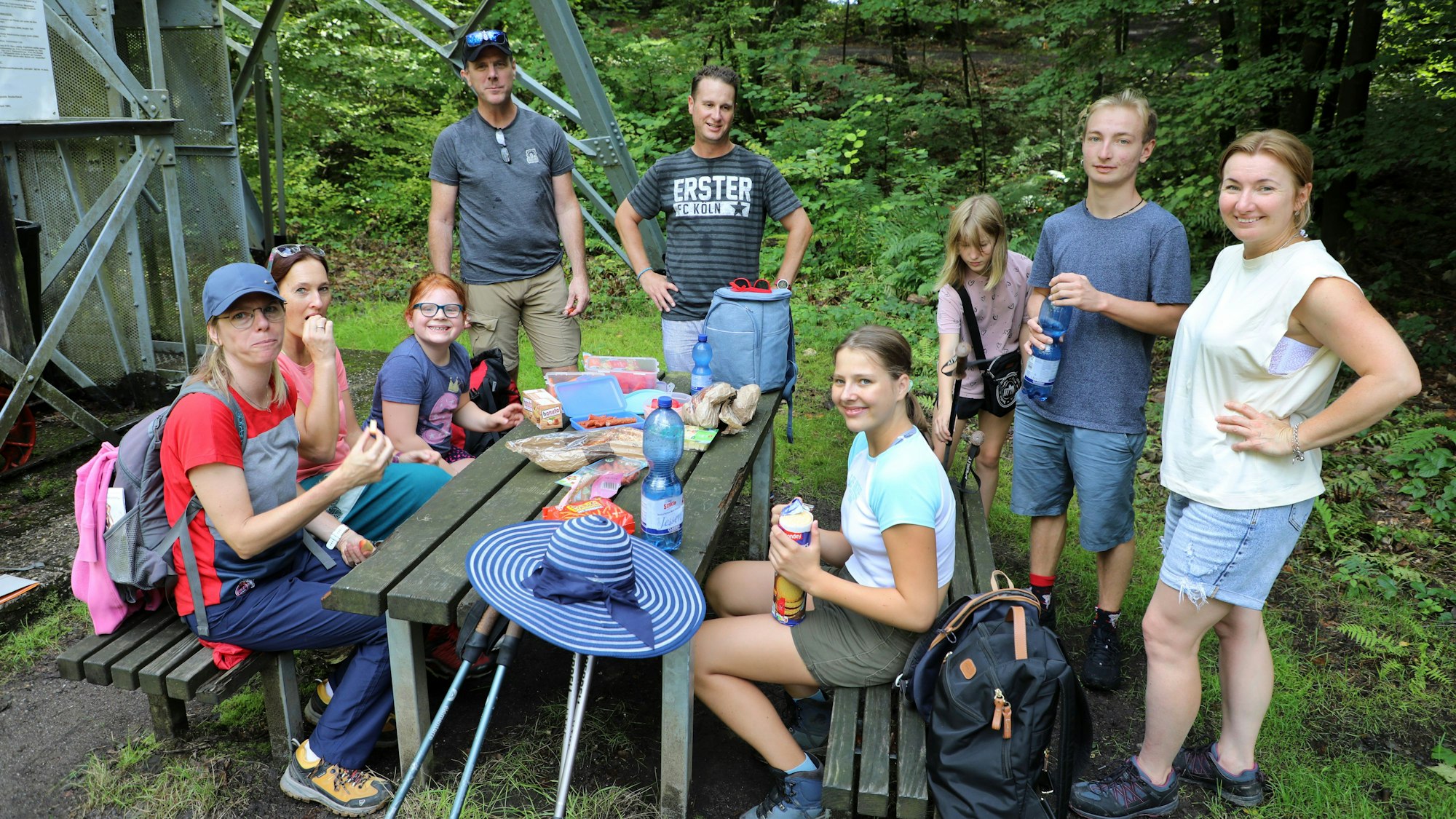 Drei Familien sitzen und stehen um einen Tisch unter freiem Himmel und machen Picknick.