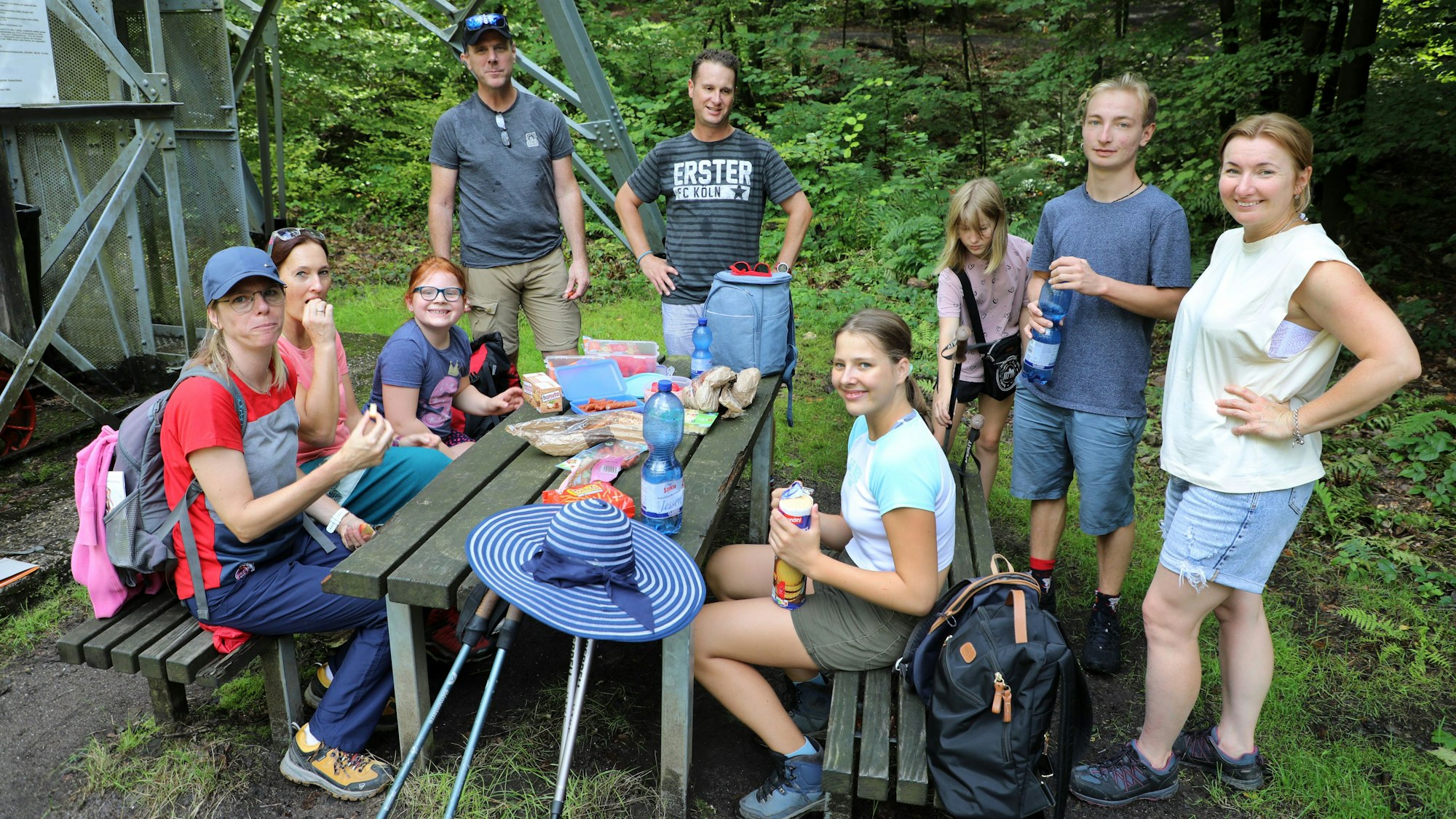 Kinder und Erwachsene sitzen und stehen an einer Tisch-Bänke-Garnitur und machen Picknick.